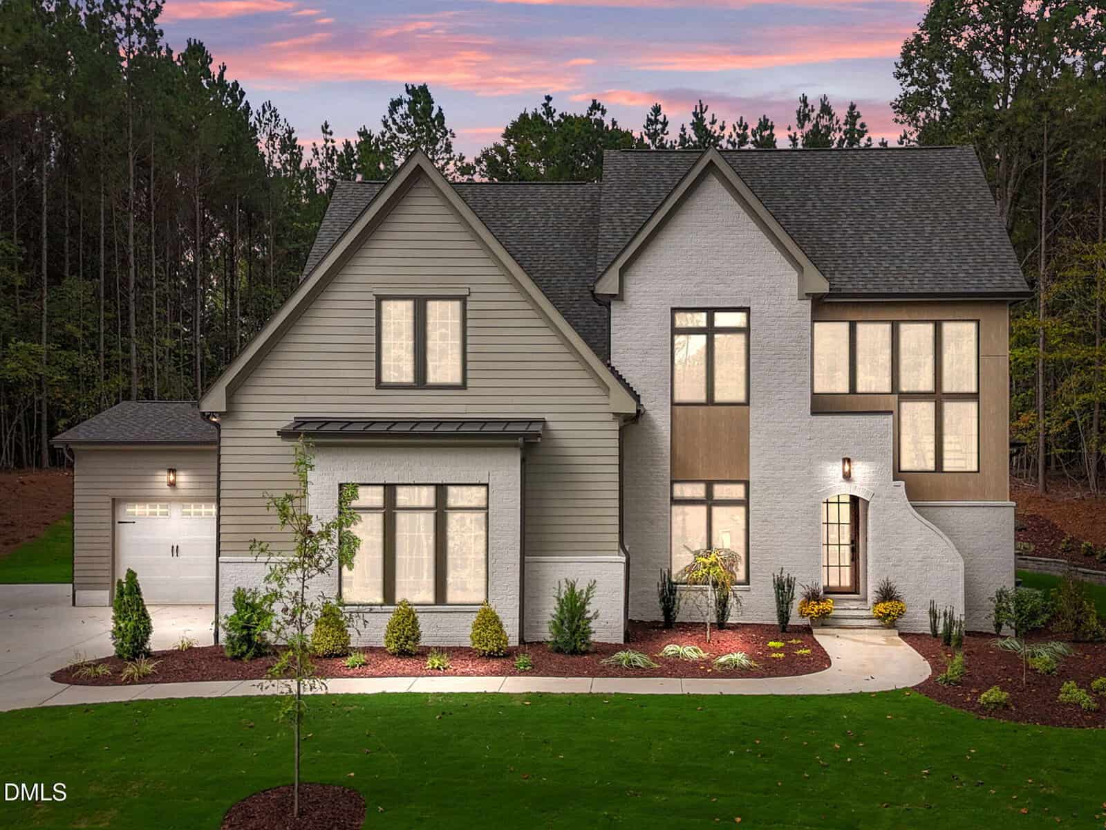 Modern two-story house at 125 Spanish Oak Drive in Youngsville, featuring beige siding with white brick accents, large windows, a two-car garage, landscaped yard, and outdoor lighting set against trees at sunset.