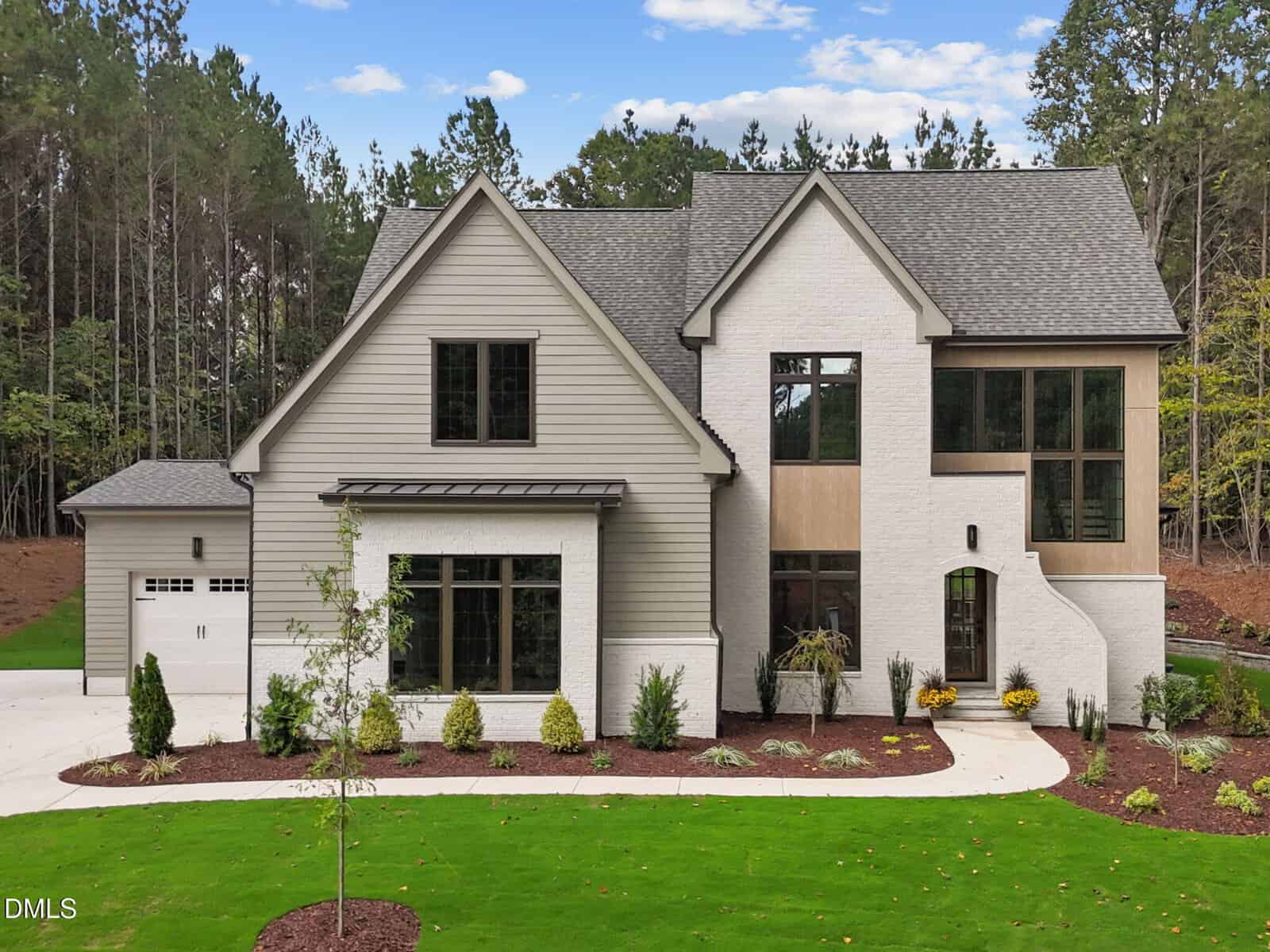 Two-story modern house with gray and white exterior, large windows, attached garage on the left, landscaped front yard with grass, small plants, and curved walkway, surrounded by tall trees and blue sky.