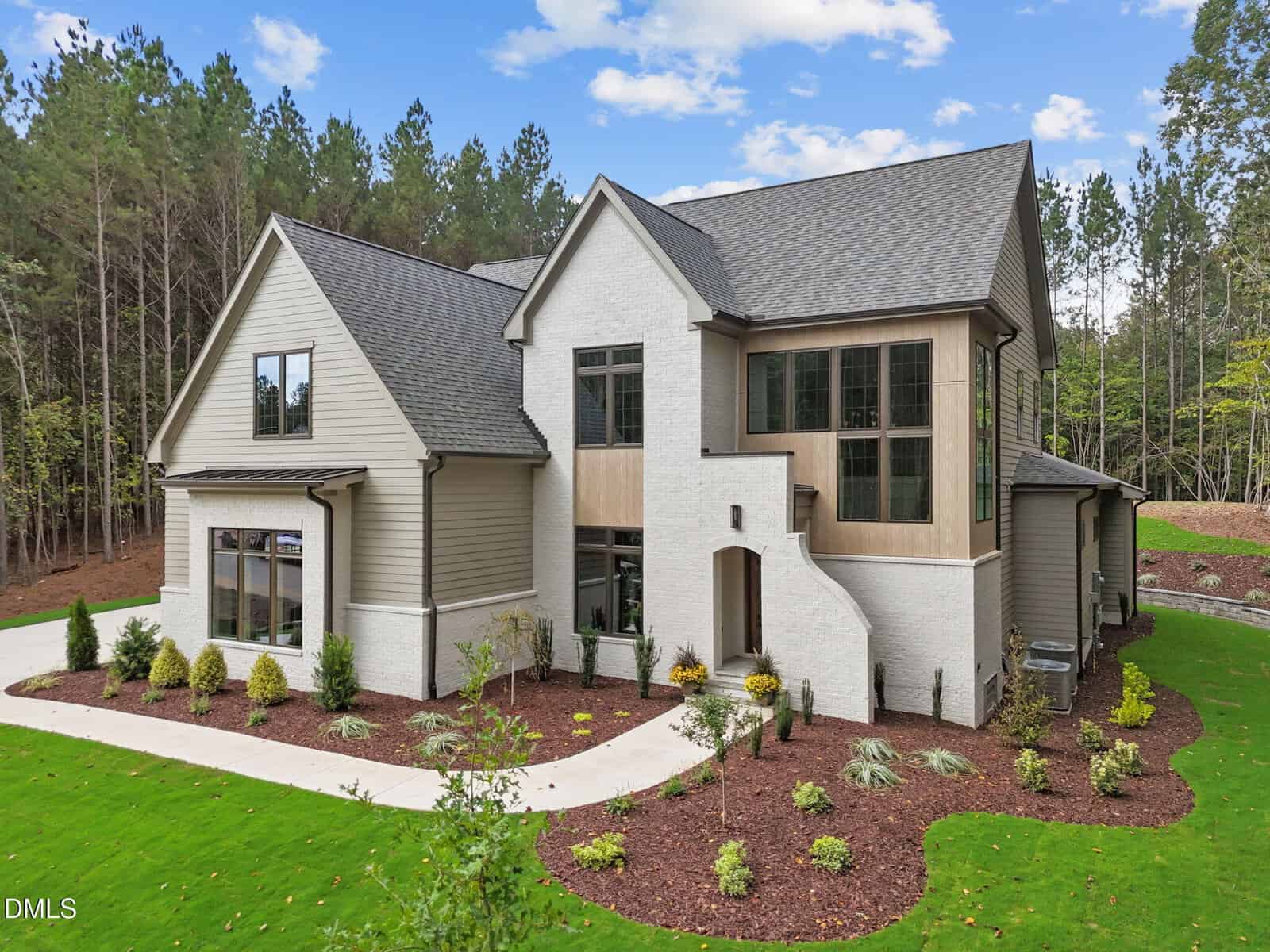 A modern two-story house with light brick, beige siding, large windows, and a landscaped yard, surrounded by trees under a blue sky with scattered clouds.