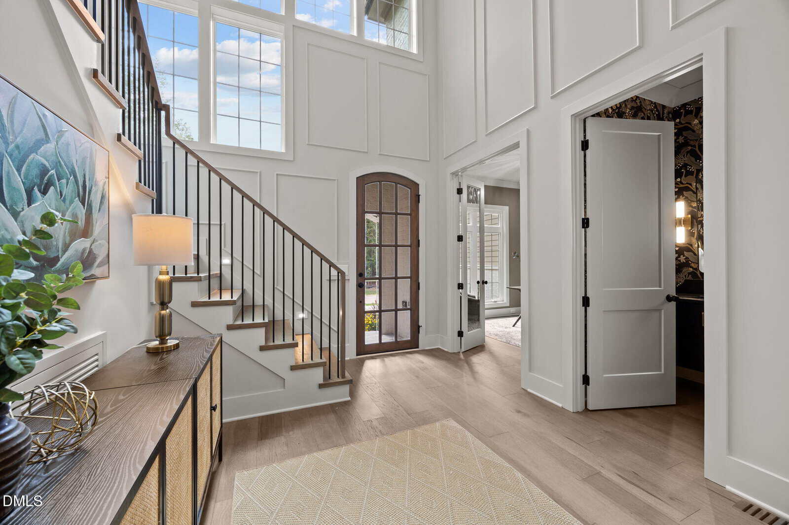 Bright entryway with light wood floors, a staircase with black railing, tall windows, a glass-paneled wooden door, a sideboard with decor, and open doors leading to other rooms. Natural light fills the space.