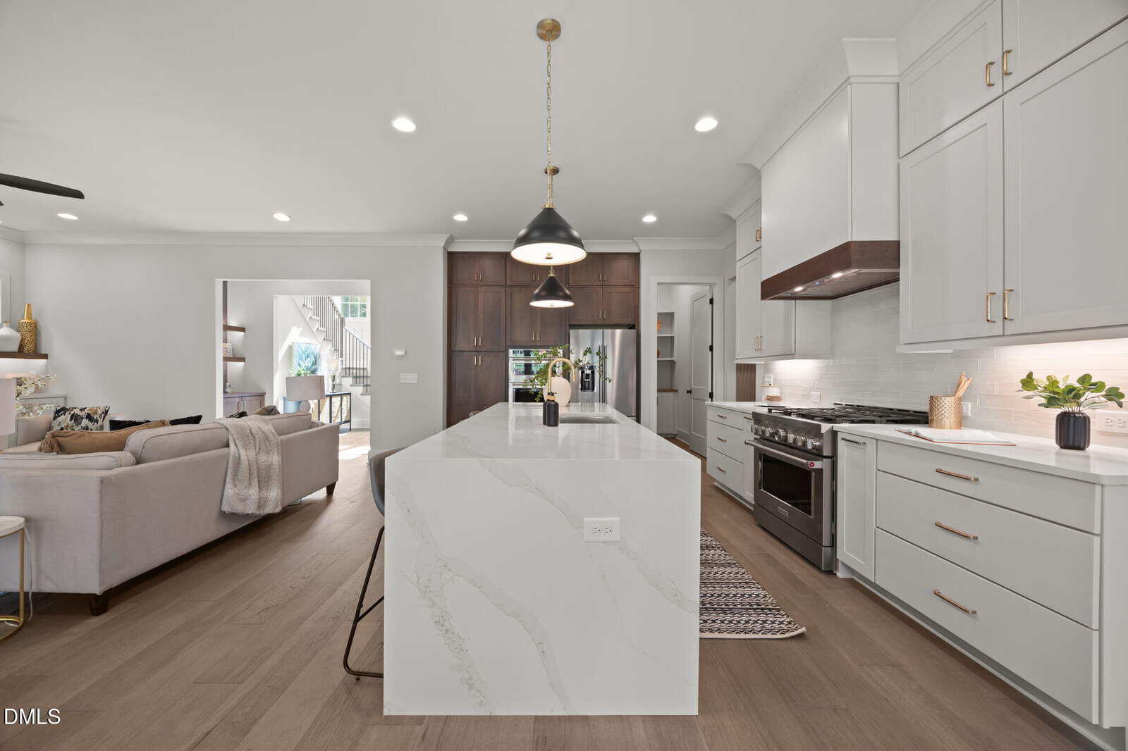 Modern kitchen with a large marble island, white cabinets, stainless steel appliances, pendant lighting, and wood flooring. An open living room with a beige sofa is visible in the background.