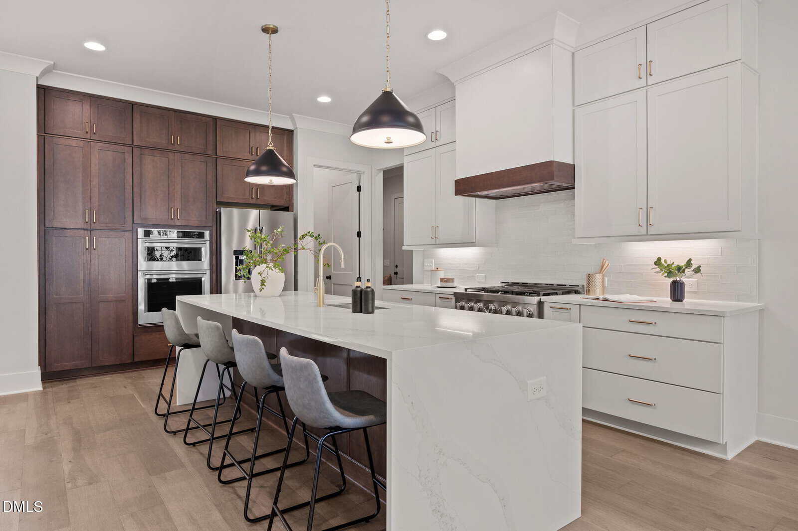 Modern kitchen with white cabinets, a large marble island with four gray barstools, stainless steel appliances, two black pendant lights, and wooden floor; dark wood cabinets and a built-in oven are in the background.