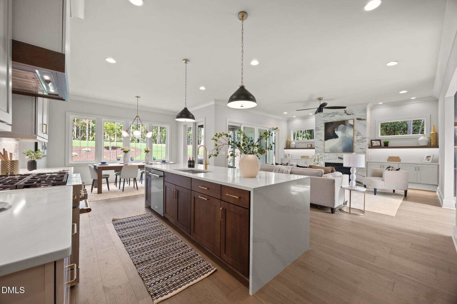 Modern open-concept kitchen and living area with a large island, marble countertops, wooden cabinets, pendant lights, dining table, and bright seating area with large windows overlooking a green backyard.