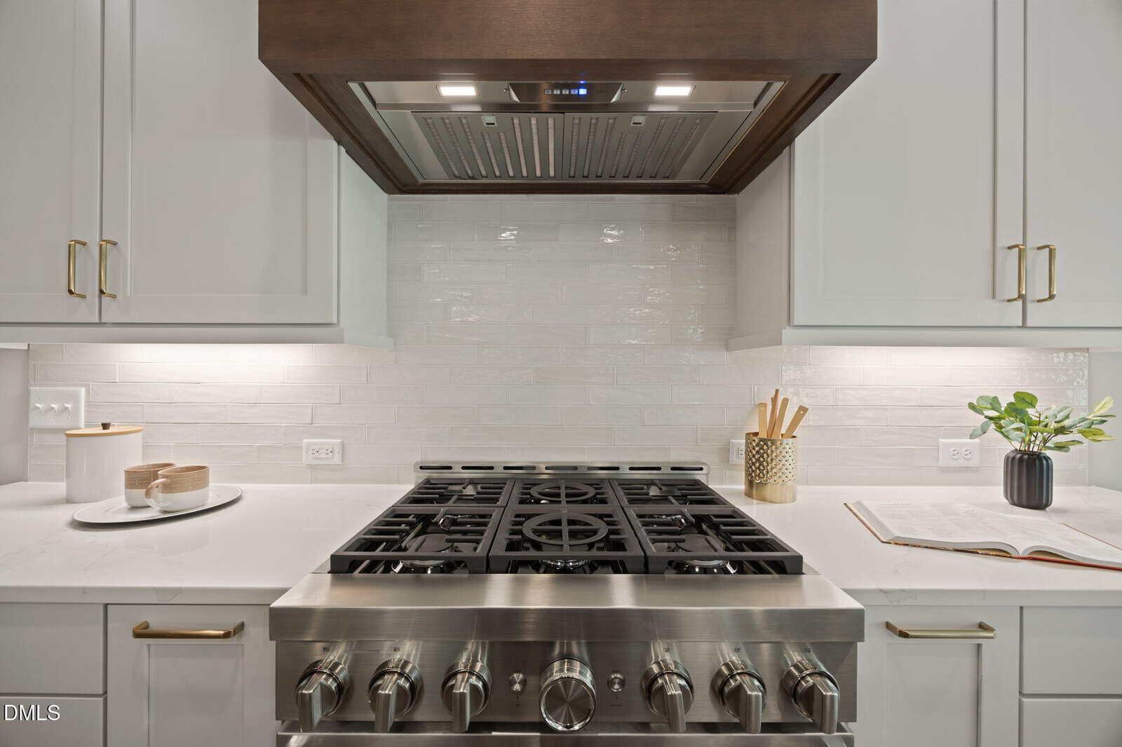 Modern kitchen with a stainless steel gas stove, white cabinets, marble countertops, utensils in a holder, an open book, a small plant, and a vent hood above the stove.