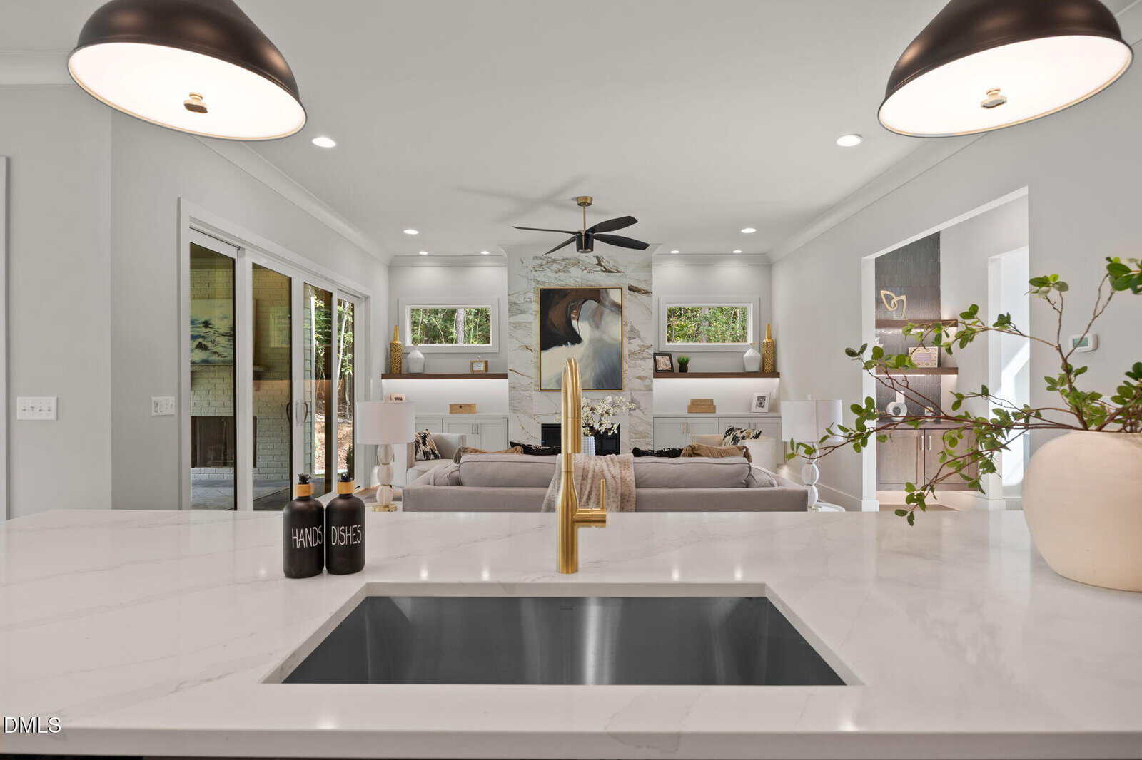 Modern kitchen island with white marble countertop and gold faucet overlooks a stylish living room with a large gray sofa, abstract art, built-in shelving, and a ceiling fan. Two pendant lights hang above the island.