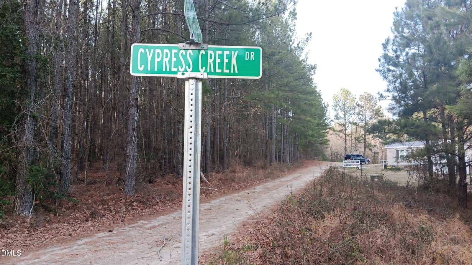 A green street sign reading “Cypress Creek Dr” stands beside a dirt road, bordered by tall pine trees, with a house visible in the distance on the right.