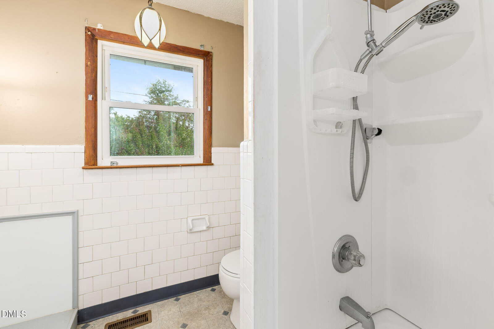 Small bathroom with white tile walls, a window with a wooden frame, a toilet, and a shower with a handheld showerhead and built-in shelves. Beige upper walls and natural light from the window.