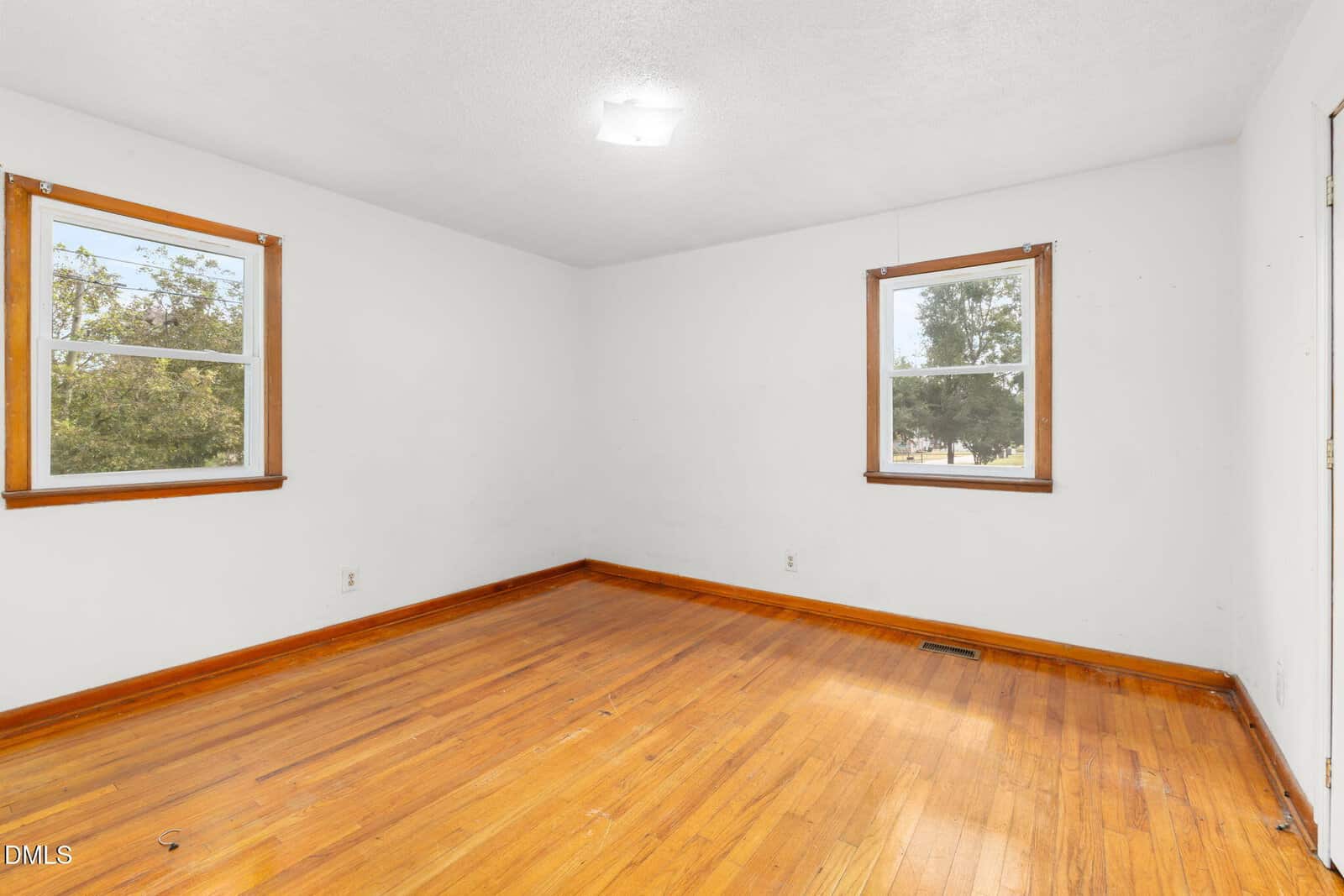 Empty room with white walls, two windows with wooden trim, hardwood floors, and a ceiling light. Trees and sky are visible outside the windows.