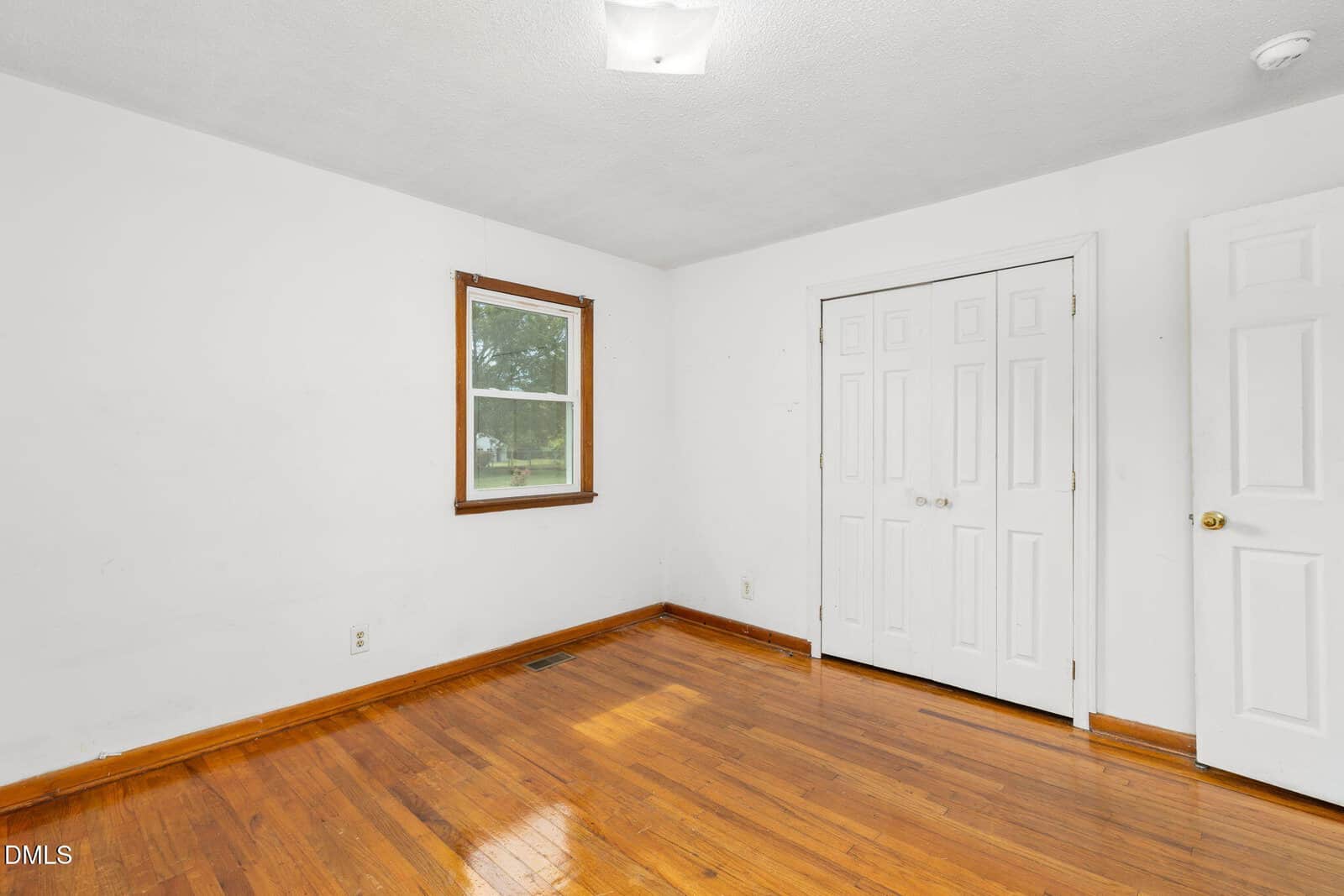 Empty room with white walls, hardwood floor, a single window, a closet with double doors, and a regular door. Ceiling light fixture is visible. Room is clean and minimally decorated.