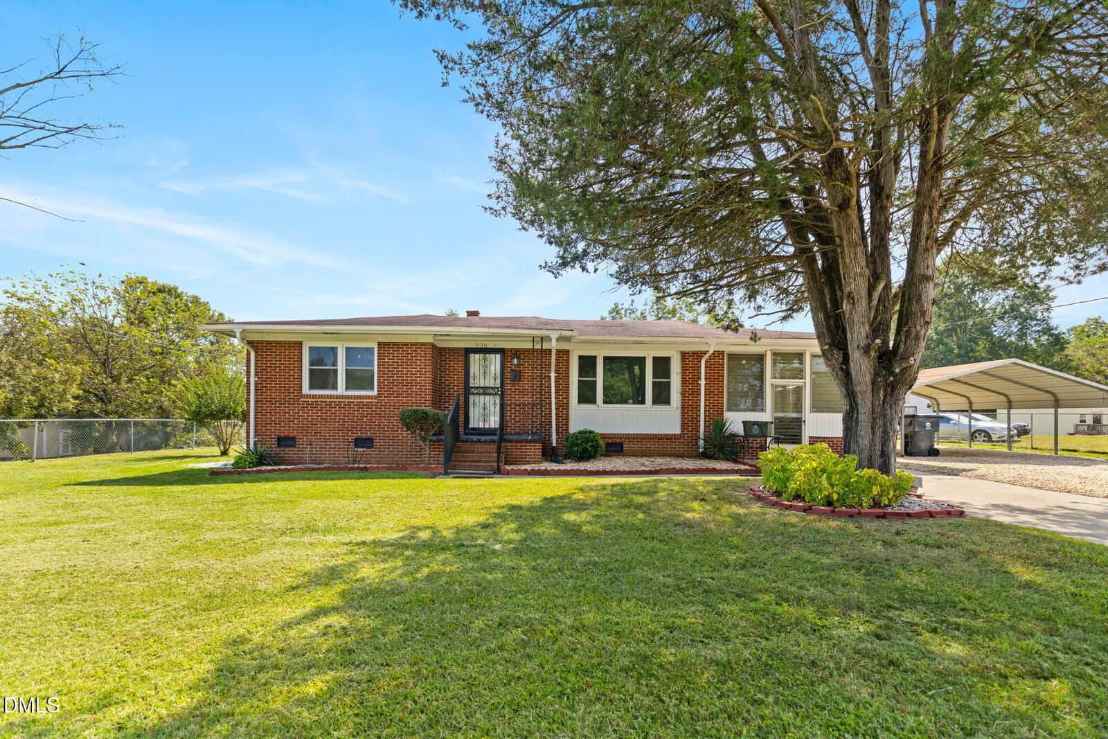 A single-story brick house with white trim, a covered front porch, landscaped bushes, a large tree in the front yard, and a driveway with a carport on the right side. The lawn is well-maintained.