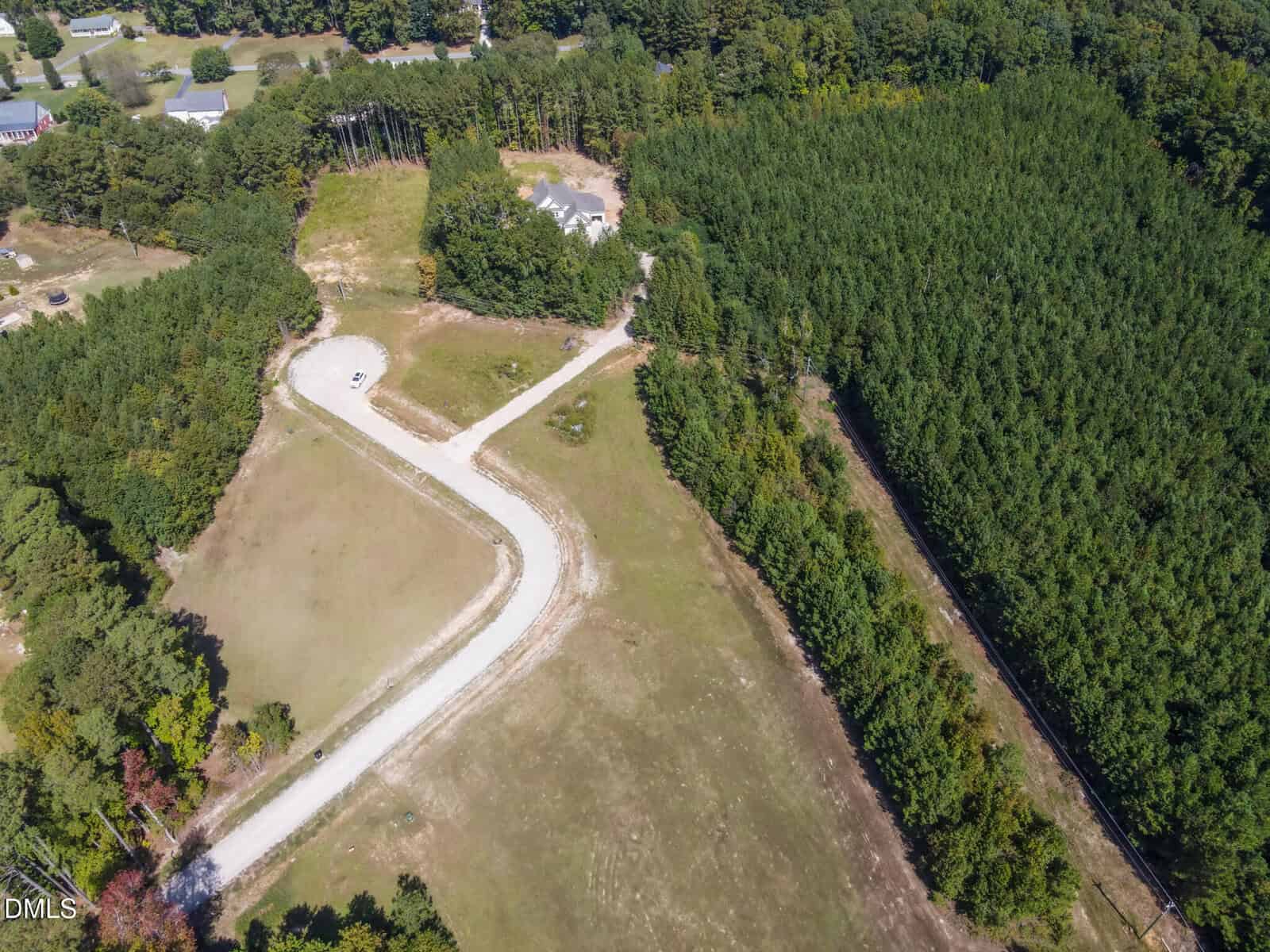 Aerial view of a rural area with a winding gravel road, a single white vehicle parked on the curve, open grassy fields, dense green forest, and a few houses scattered in the background.