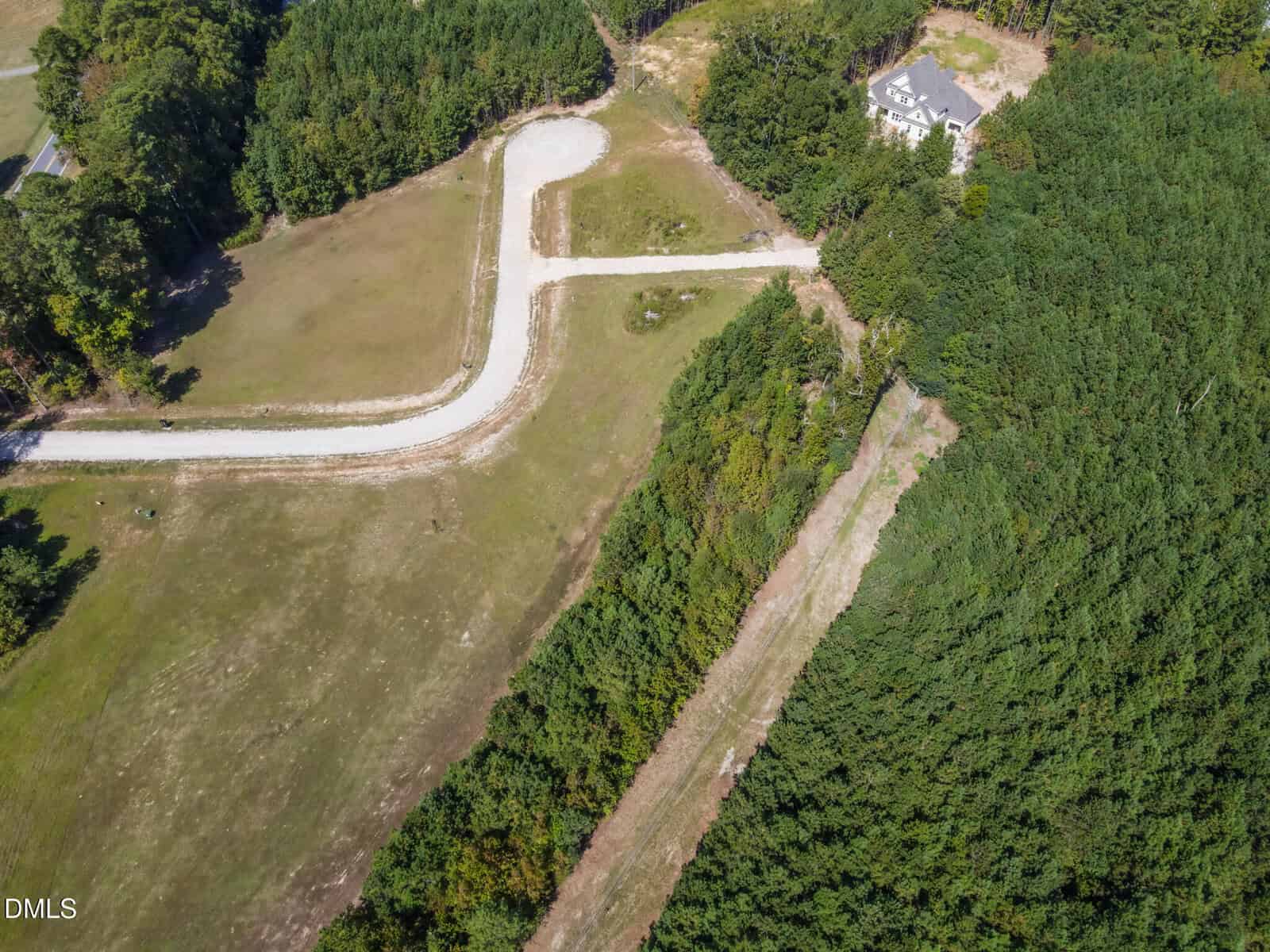 Aerial view of a curved, unpaved road leading to a cul-de-sac surrounded by trees and grassy fields, with a house partially visible among trees in the upper right corner.