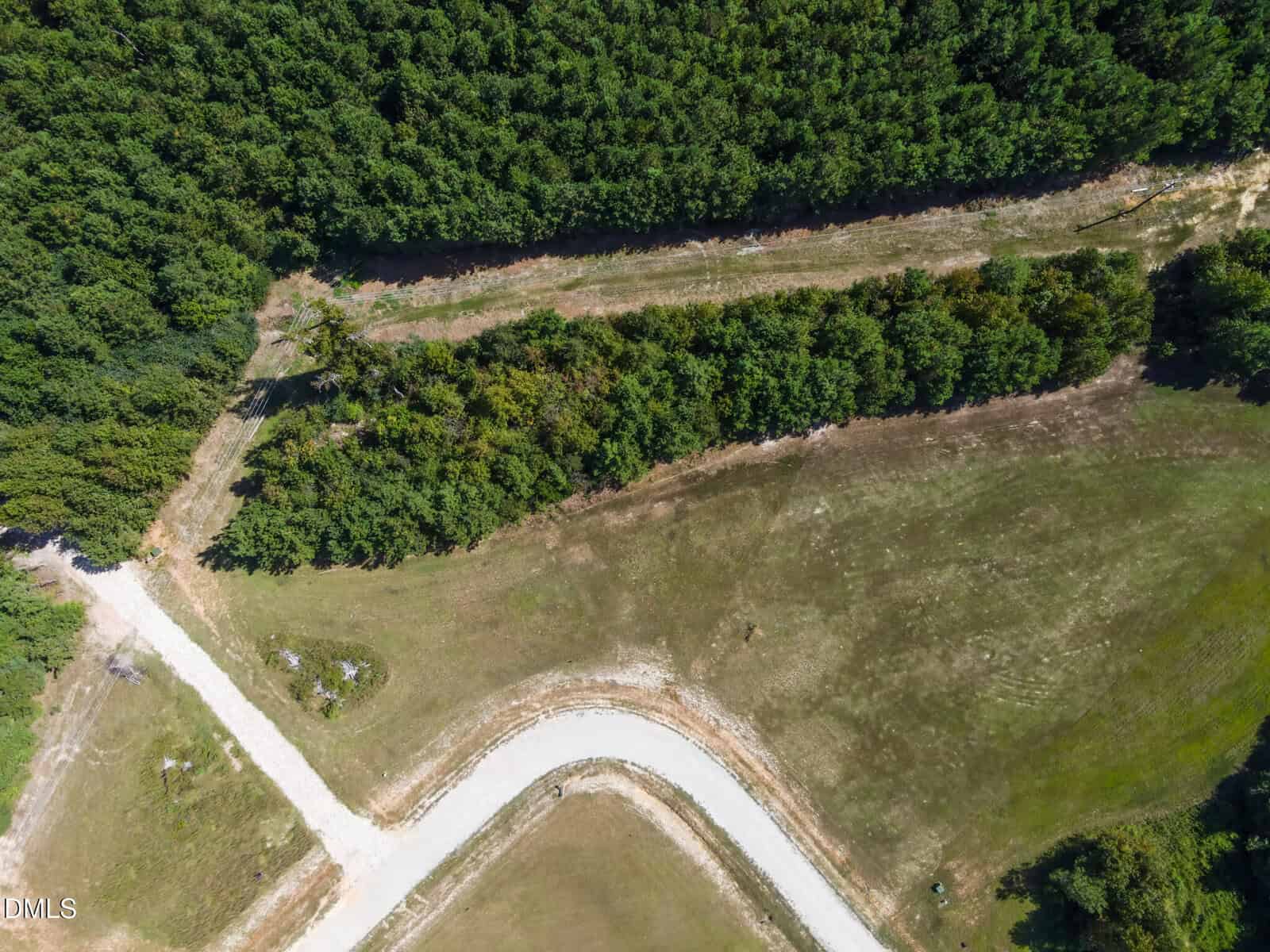 Aerial view of a curved, paved road meeting a dirt path surrounded by grassy fields and dense green trees at the top and left side of the image.