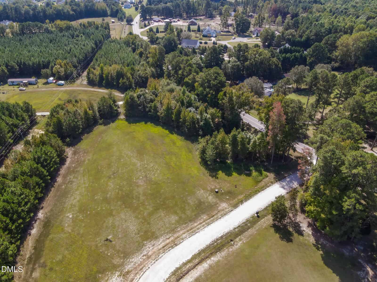 Aerial view of a rural area with a winding gravel road leading through open grassy land, surrounded by dense green trees, and a small town visible in the background.