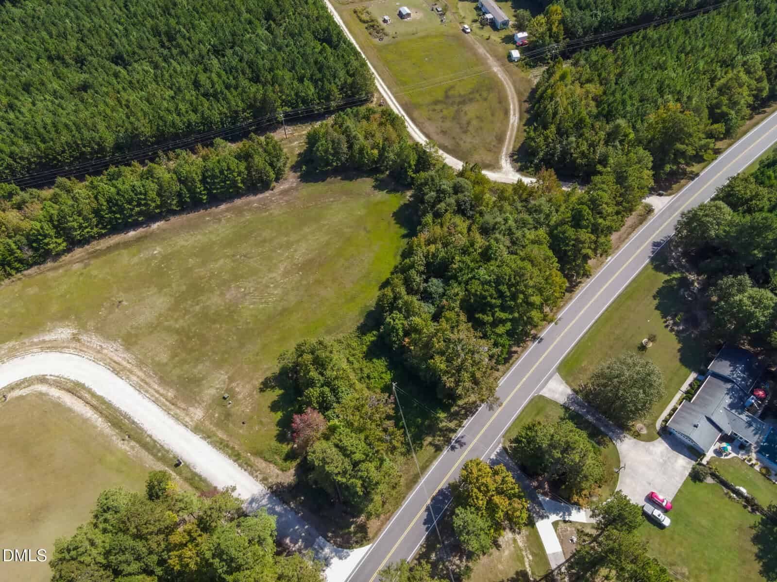 Aerial view of a rural area with a paved road curving through fields, surrounded by trees and houses. A smaller dirt road intersects, and open grassy spaces are bordered by dense forest.