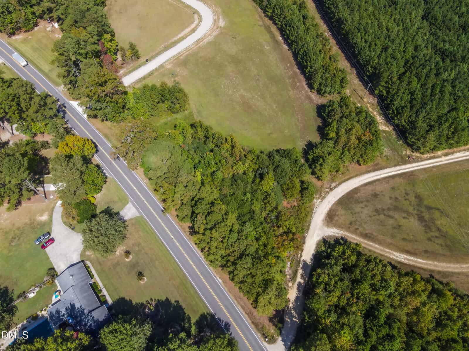 Aerial view of a rural intersection with a paved road, two curved dirt roads, a house with a driveway, and surrounding grassy fields and trees.