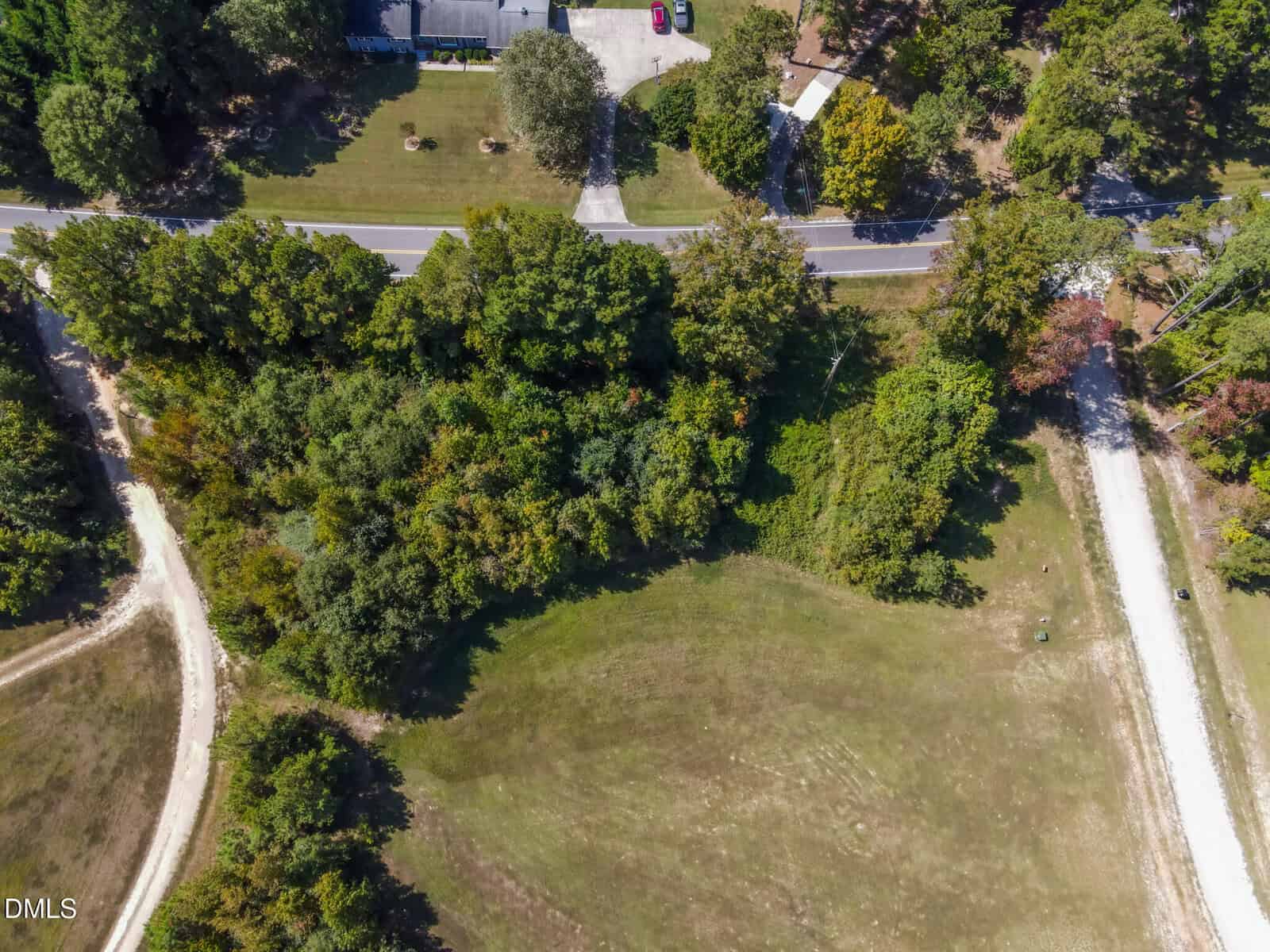 Aerial view of a grassy, partially wooded lot bordered by two dirt roads, with a paved road and houses visible near the top edge of the image. Trees and open grass areas are prominent in the scene.