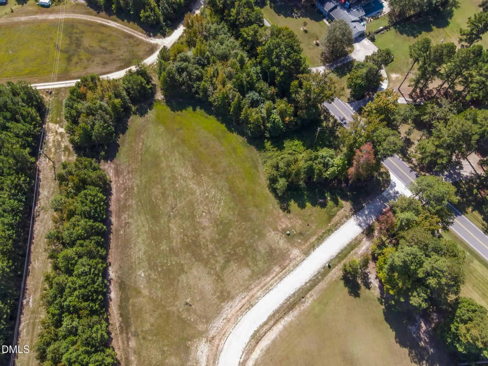 Aerial view of a grassy, partially cleared lot bordered by trees, with a curved white gravel driveway leading to a paved road. Surrounding areas have scattered trees and nearby buildings.