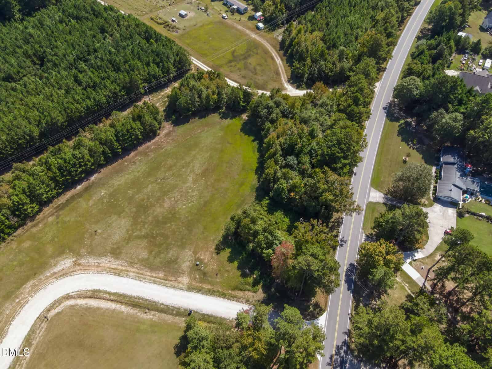 Aerial view of a rural area with a curved dirt road, grassy open field, dense green trees, and a paved road running alongside homes and wooded land.