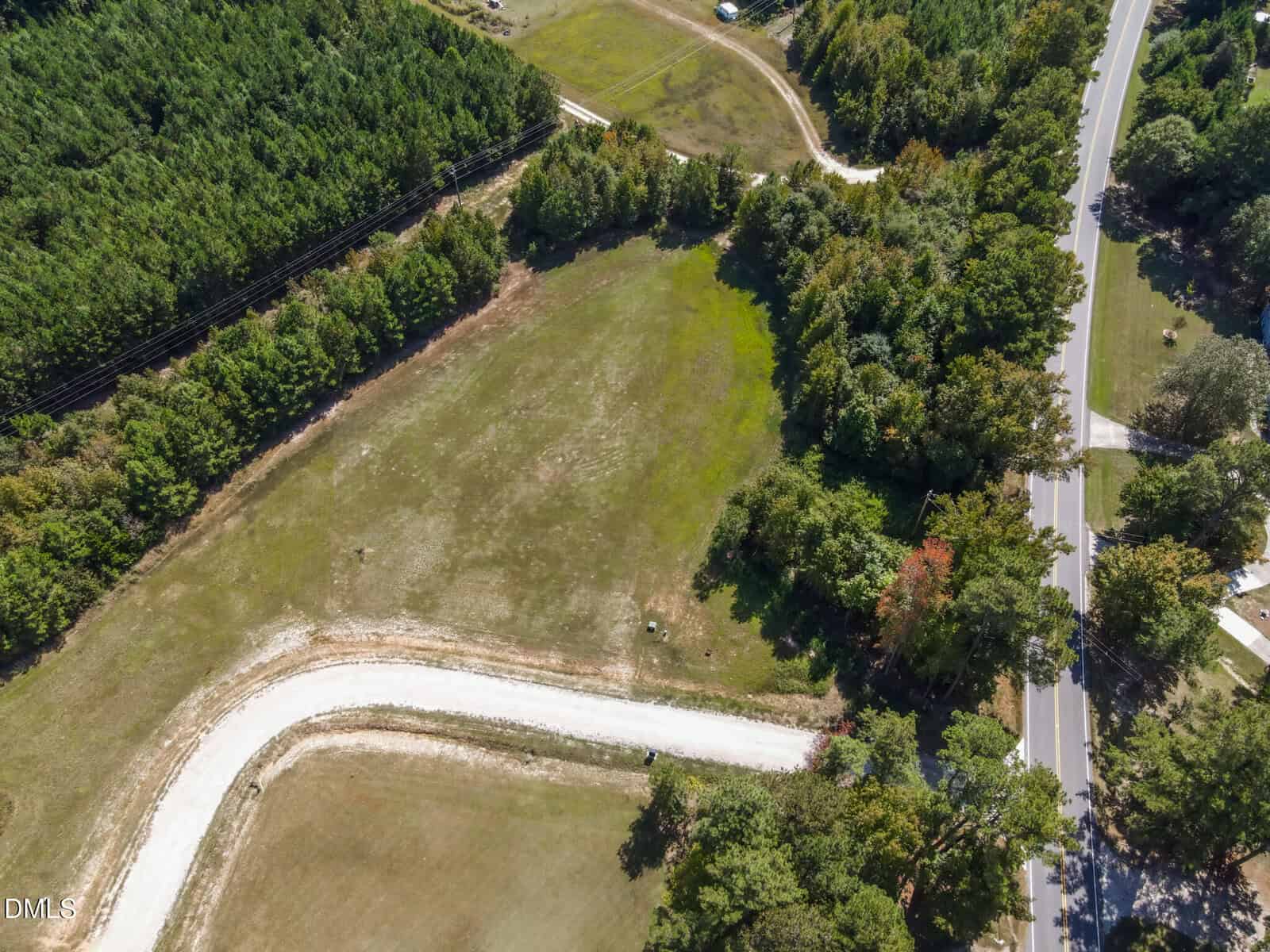 Aerial view of a cleared, grassy plot of land bordered by dense trees, with a curved gravel road and an adjacent paved road running through a wooded rural area.