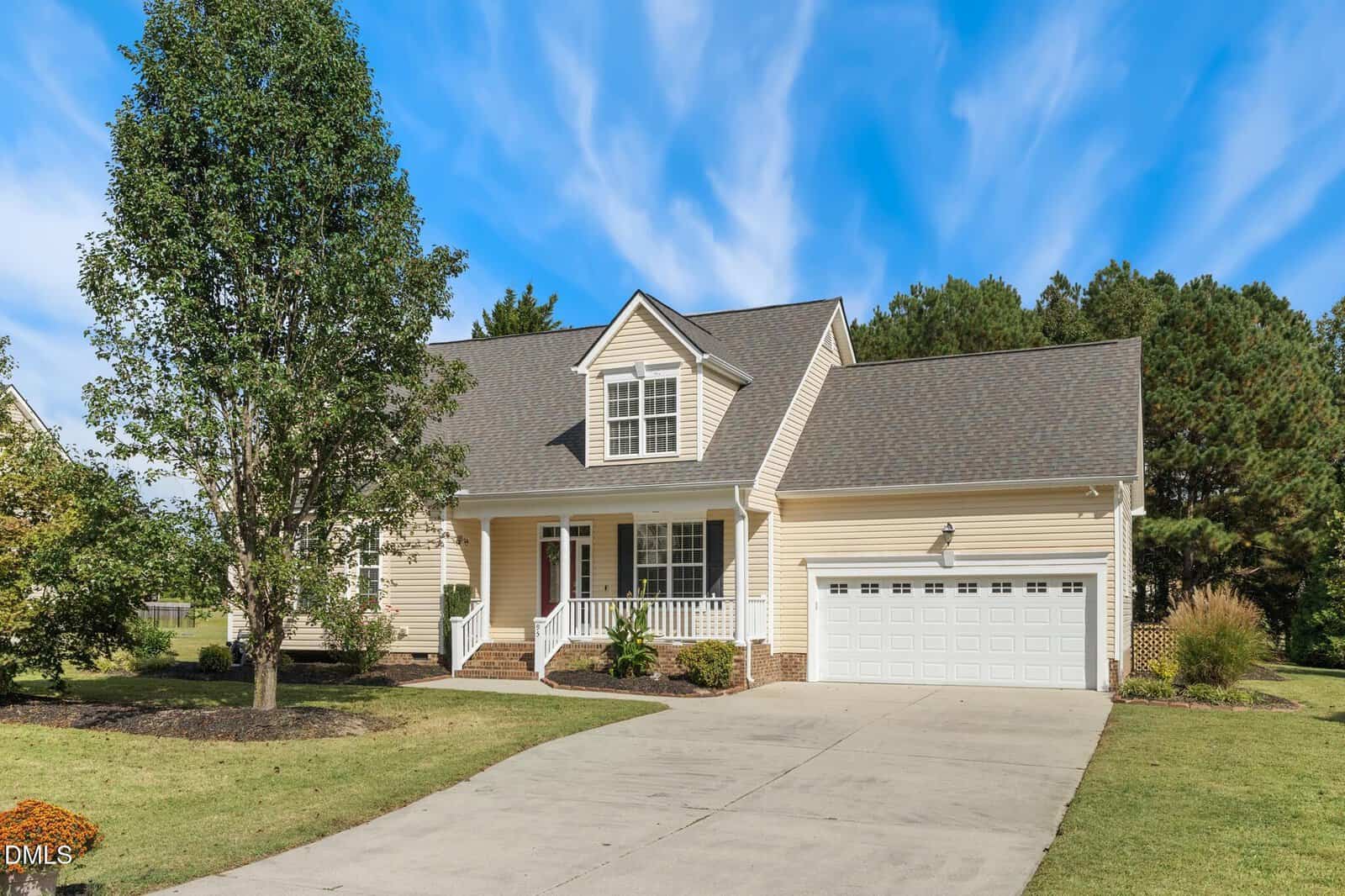 This two-story beige house with white trim at 95 Glen Oaks Drive in Youngsville features a two-car garage, a covered front porch, a large tree in the yard, and a wide driveway under a blue sky with wispy clouds.
