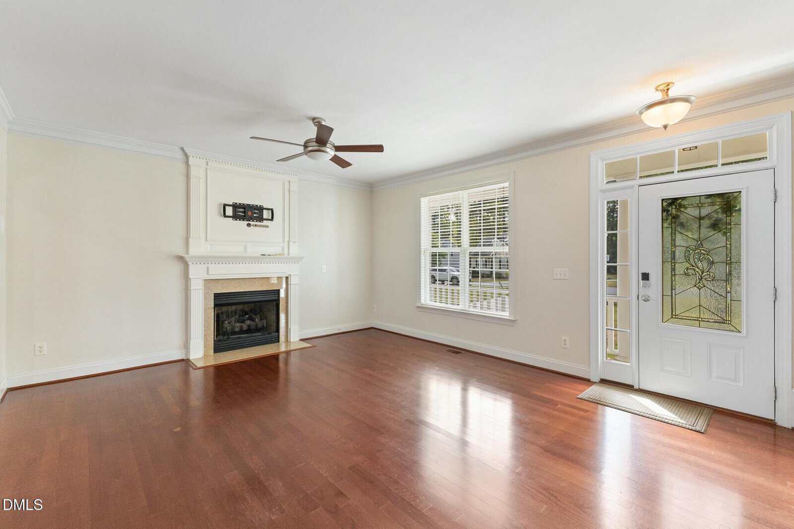 A bright, empty living room with wood floors, a white fireplace and mantel, a ceiling fan, large windows, and a white front door with decorative glass panels.