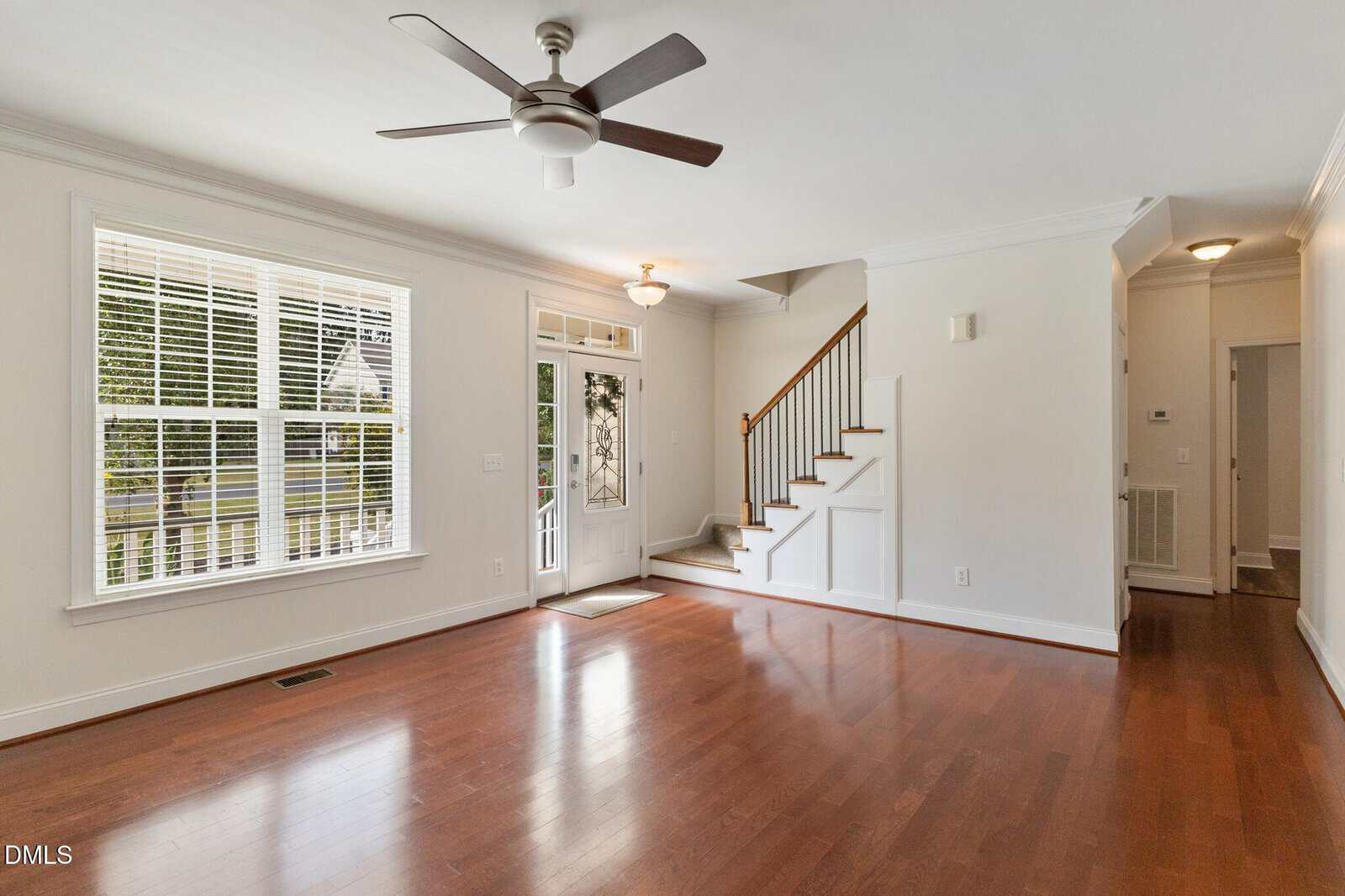 Bright, empty living room with hardwood floors, a large window, ceiling fan, white walls, and a staircase leading upstairs near the front door. Natural light enters through the window and glass door.