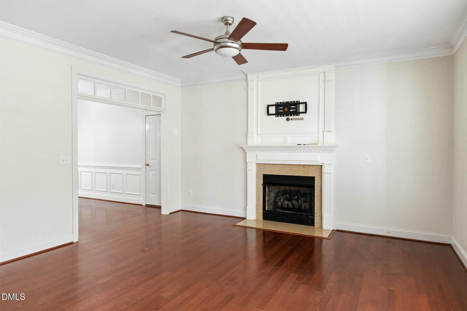 A living room with hardwood floors, neutral walls, a ceiling fan, and a fireplace with a mounted TV above it. An adjacent room with wainscoting is visible through an open doorway.