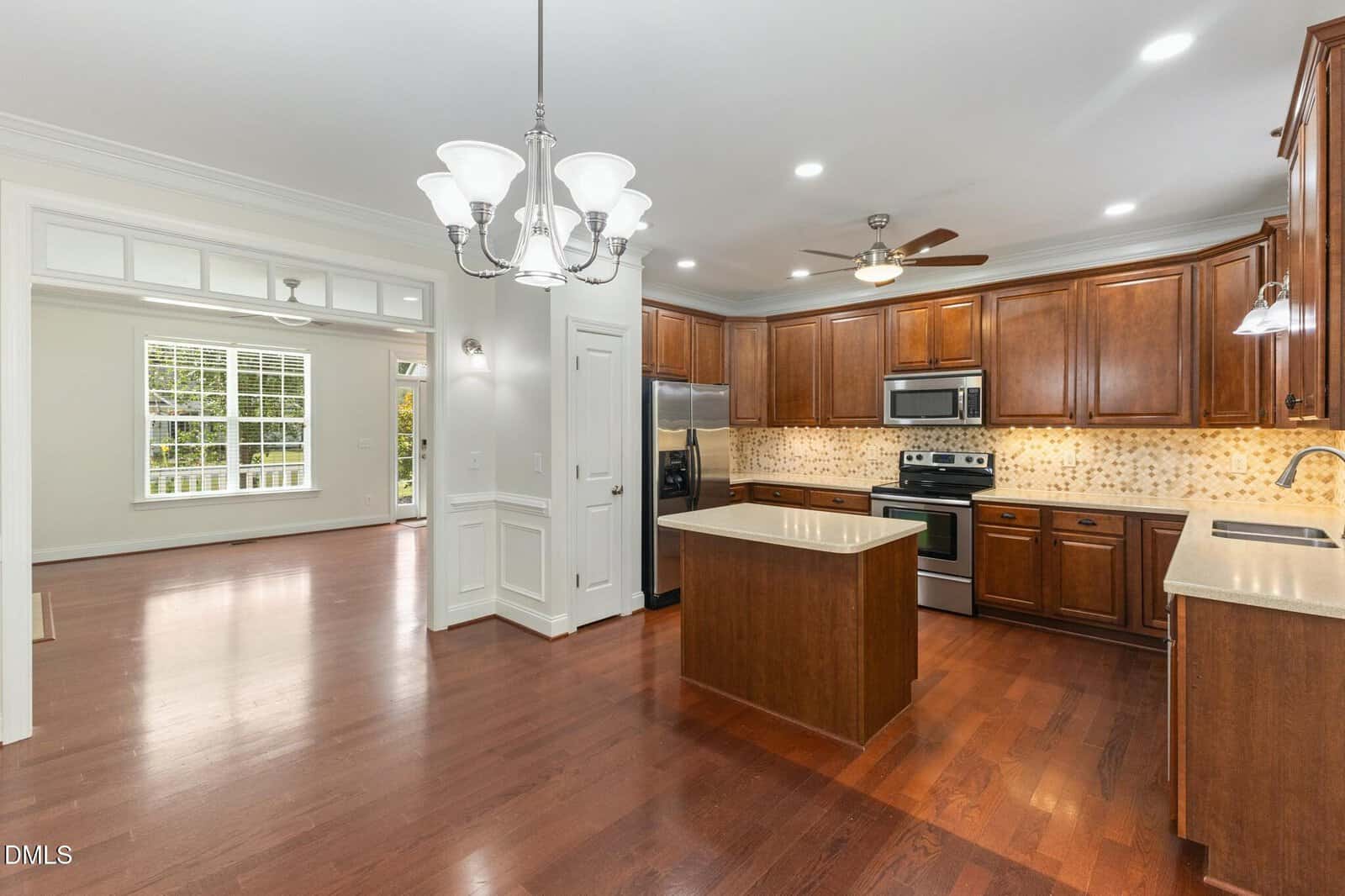 Spacious kitchen with wooden cabinets, island, stainless steel appliances, and mosaic backsplash. Open layout leads to a bright living area with large windows and hardwood floors throughout.