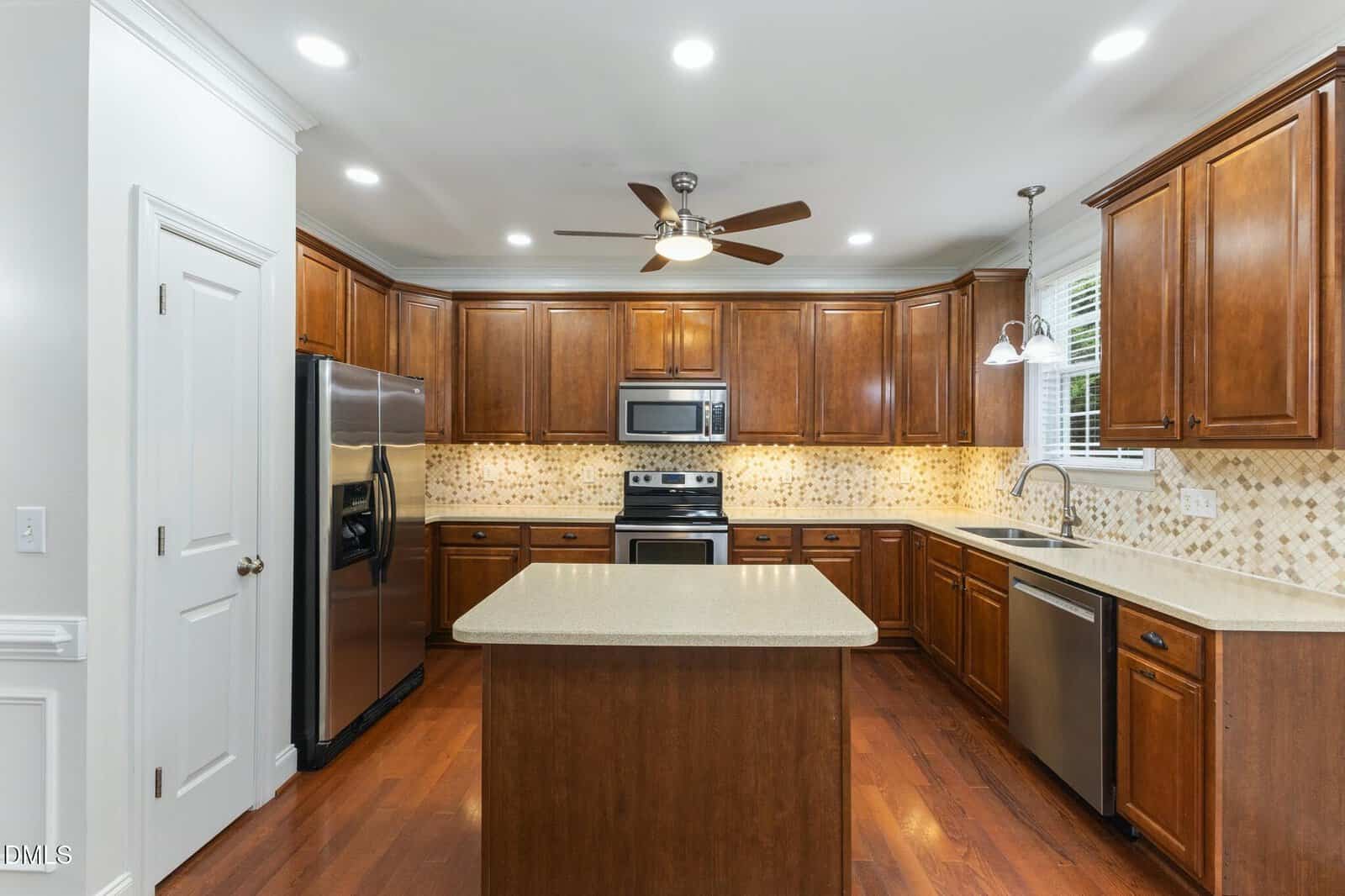 Modern kitchen with wood cabinets, stainless steel appliances, a central island, tile backsplash, ceiling fan, and recessed lighting. There is a window above the sink and hardwood floors throughout.