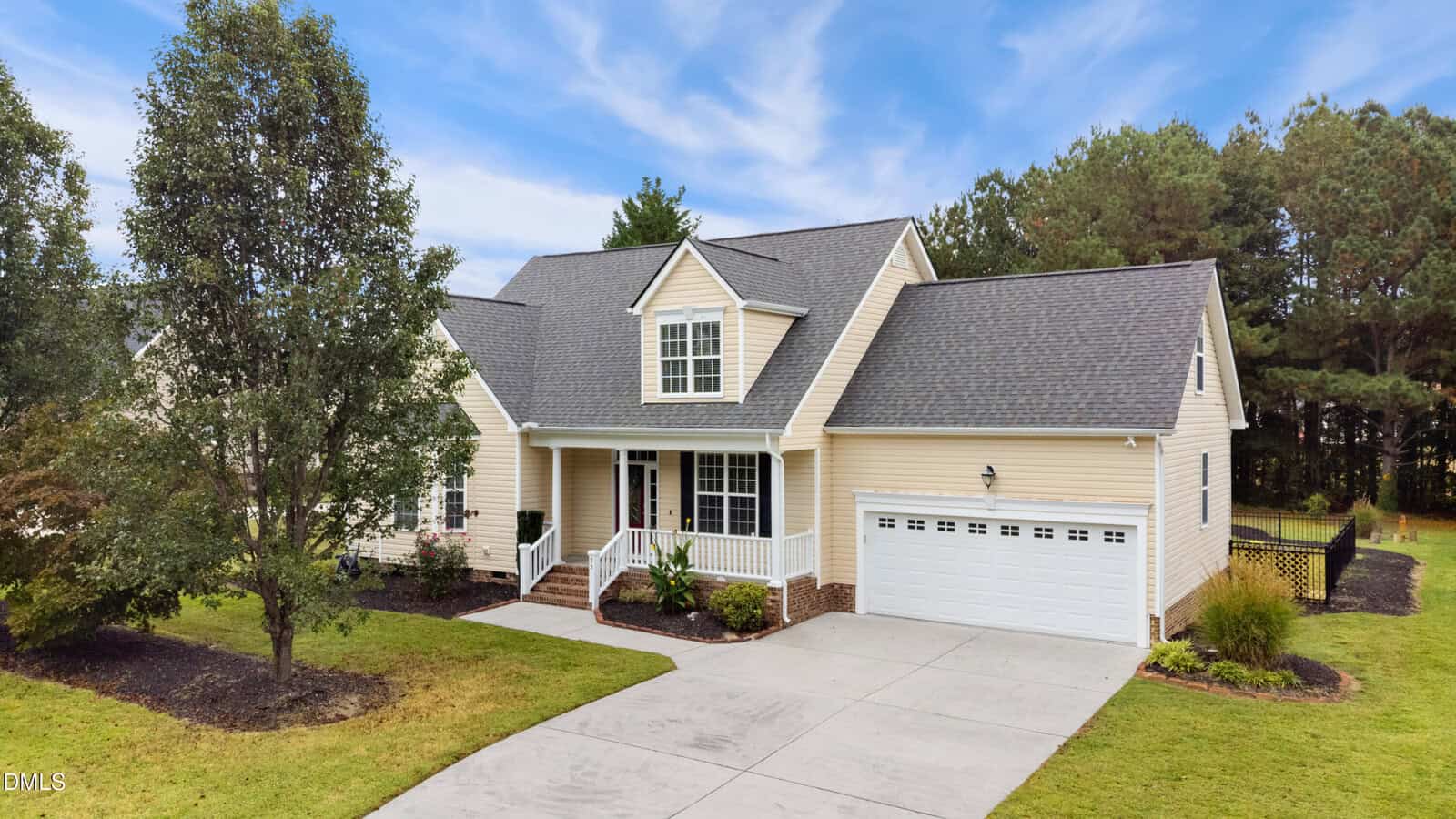 A two-story beige house with a covered front porch, white trim, and an attached two-car garage. The house is surrounded by a lawn, landscaping, and trees, with a wide concrete driveway leading to the garage.