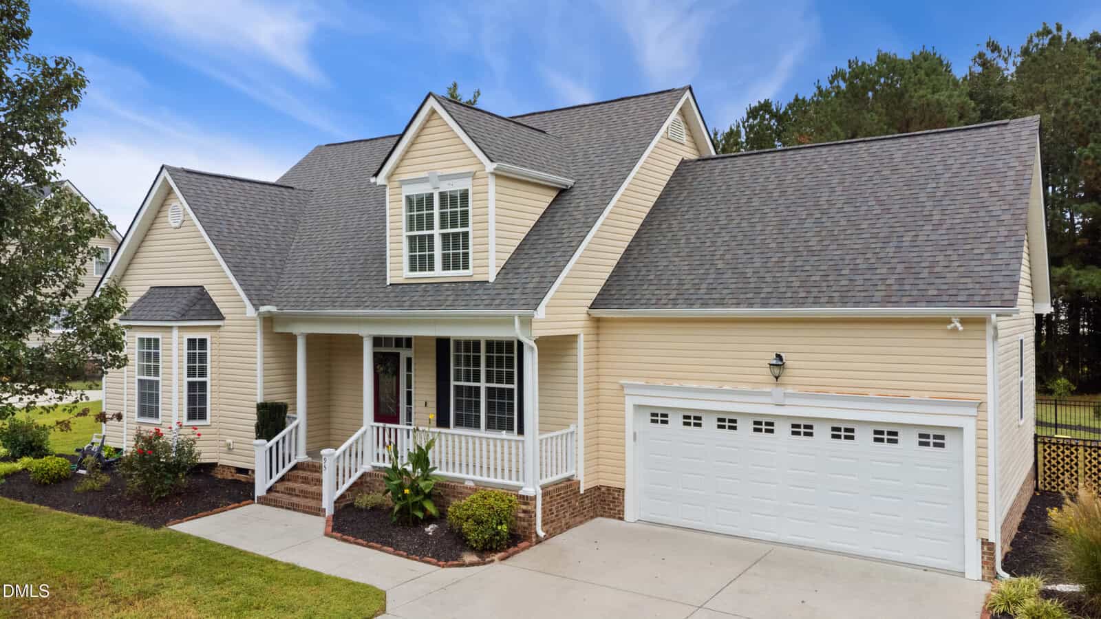 A beige, two-story suburban house with white trim, a covered front porch, and an attached two-car garage. There are landscaped bushes and a small lawn in front, with a backdrop of trees and a blue sky.