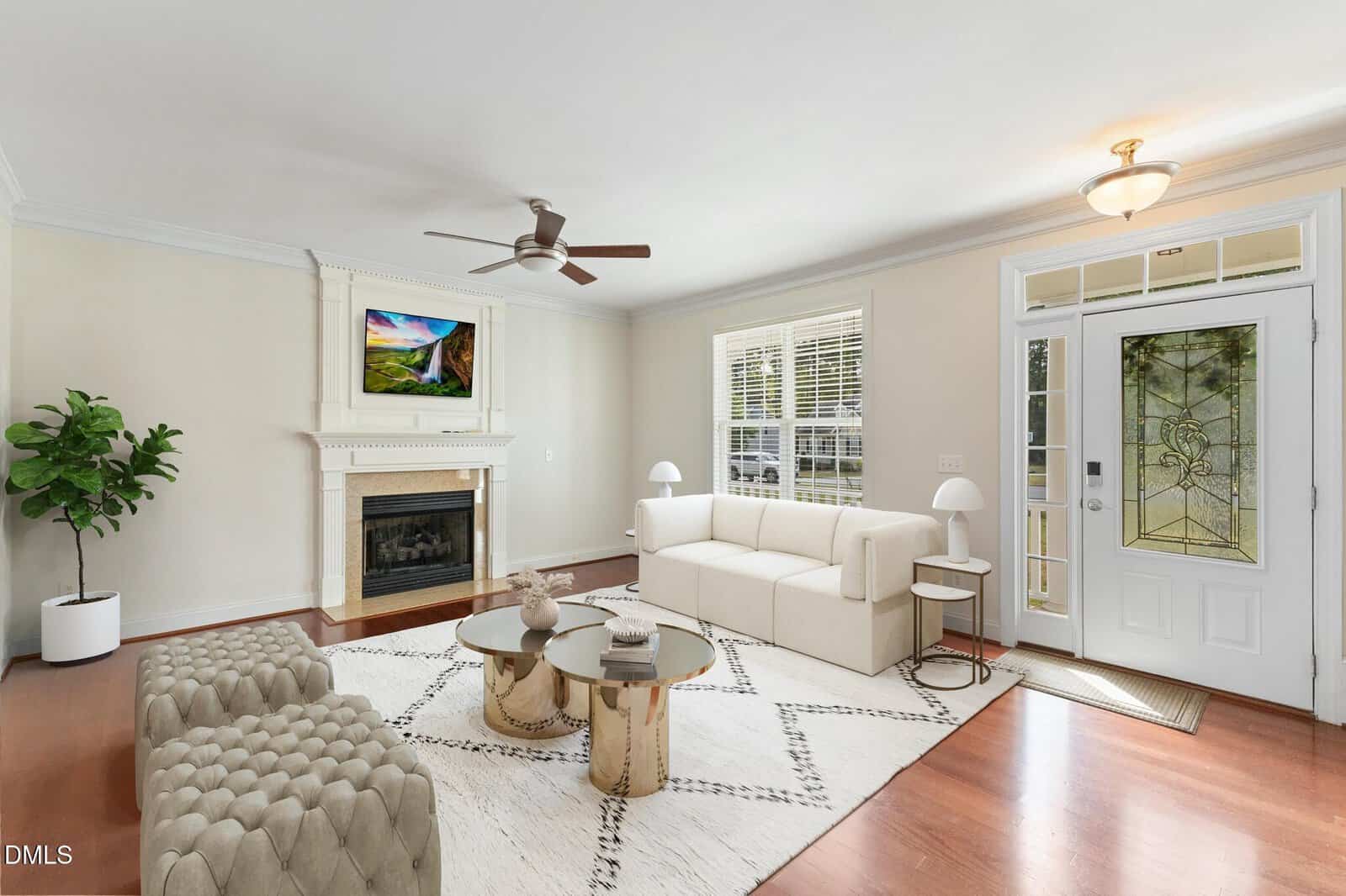 A bright living room with a beige sectional sofa, two tufted ottomans, glass coffee tables, a fireplace with a TV above it, a potted plant, and a large window near the white front door with glass panels.
