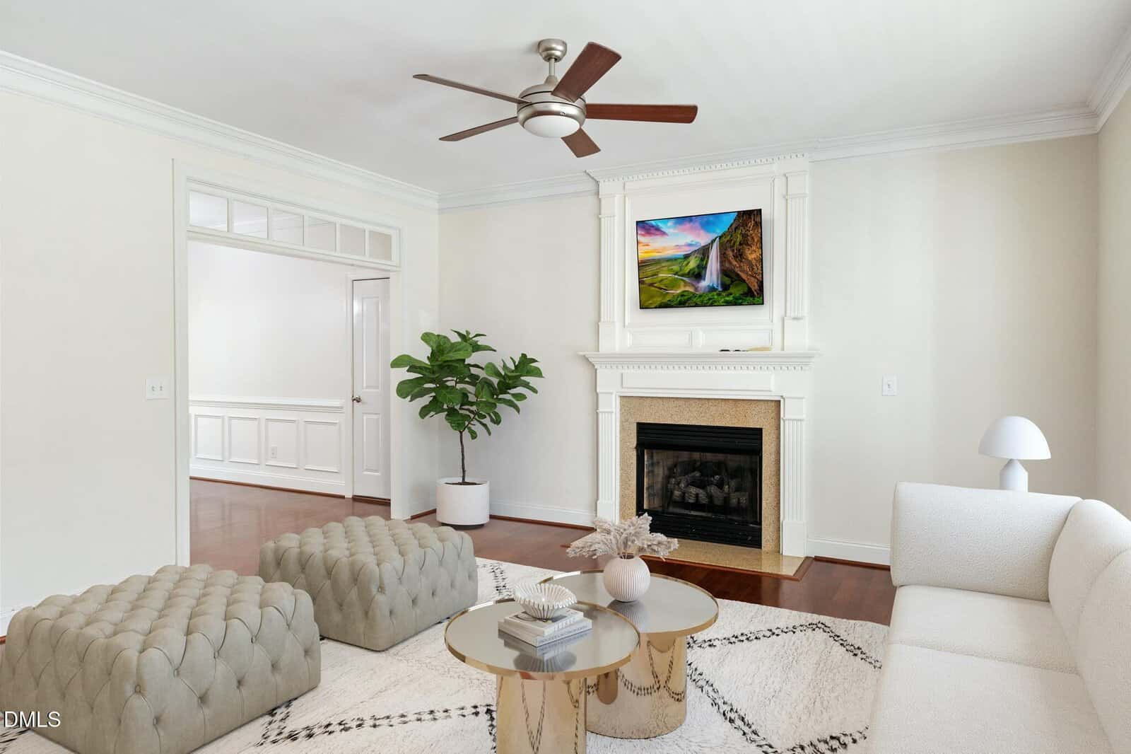 A living room with a white sofa, two gray ottomans, a round glass coffee table, a fireplace with a TV above it, a potted plant, and white walls with crown molding.