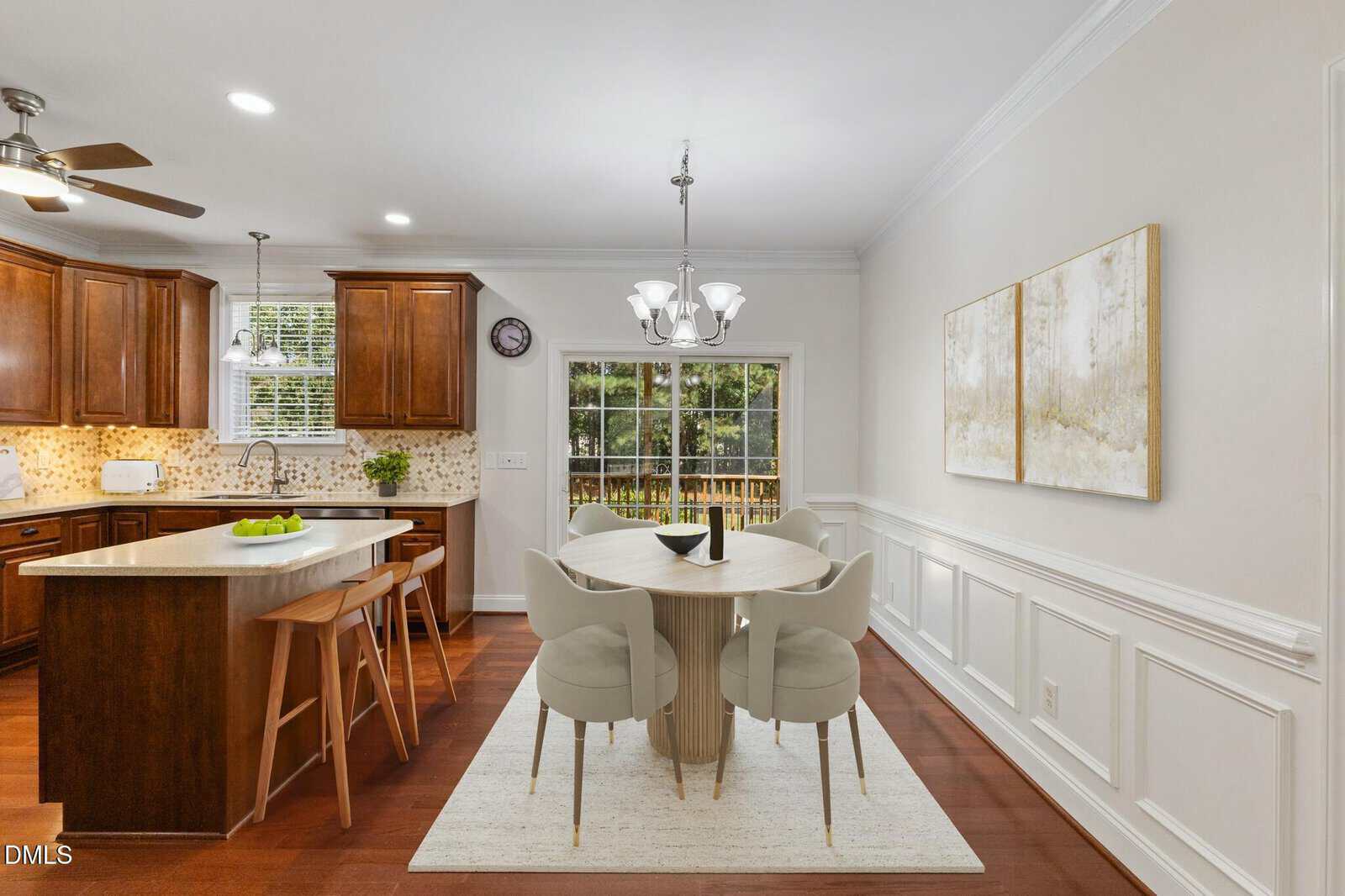 A kitchen and dining area with wooden cabinets, a central island, a round dining table with four chairs on a rug, two wall art pieces, and large glass doors overlooking a backyard with a wooden fence.