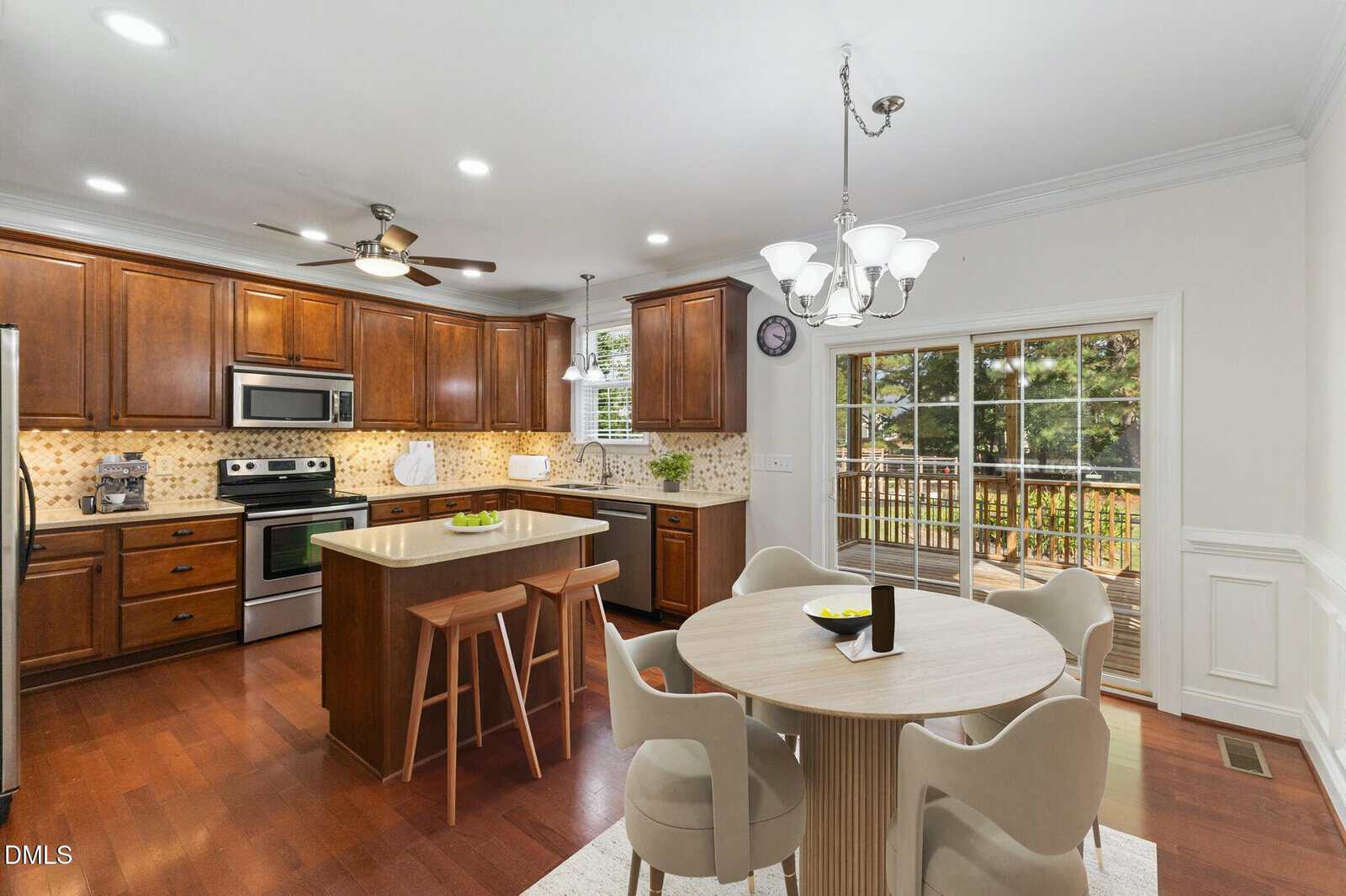 A kitchen with wooden cabinets, stainless steel appliances, an island with two stools, and a round dining table with four chairs. Sliding glass doors open to a deck outside. The room is well-lit with ceiling and wall lights.