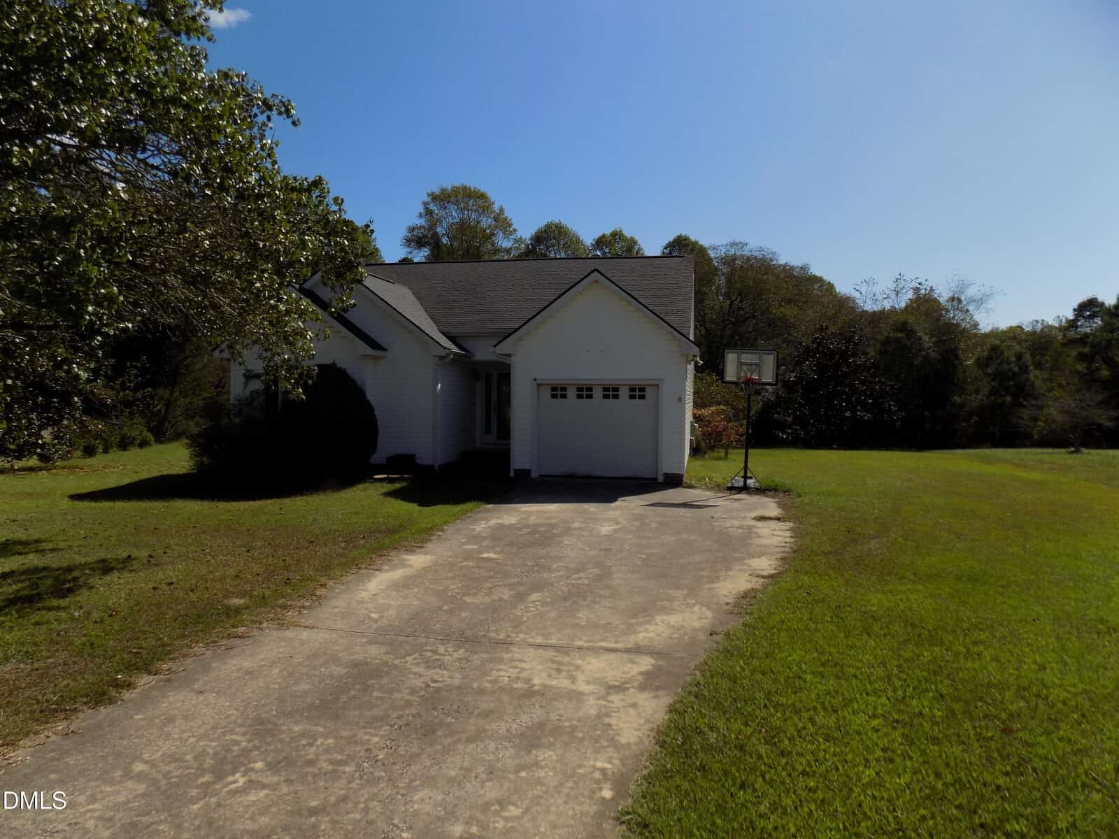A small white house with an attached single-car garage and a driveway. There is a basketball hoop by the driveway and a large yard with trees and grass. The sky is clear and sunny.