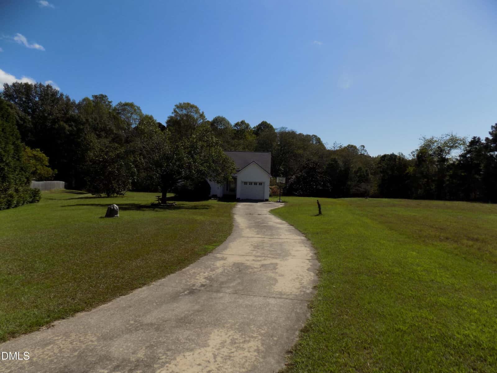 A long curved concrete driveway leads to a small white house with a garage, surrounded by green grass and trees under a clear blue sky.