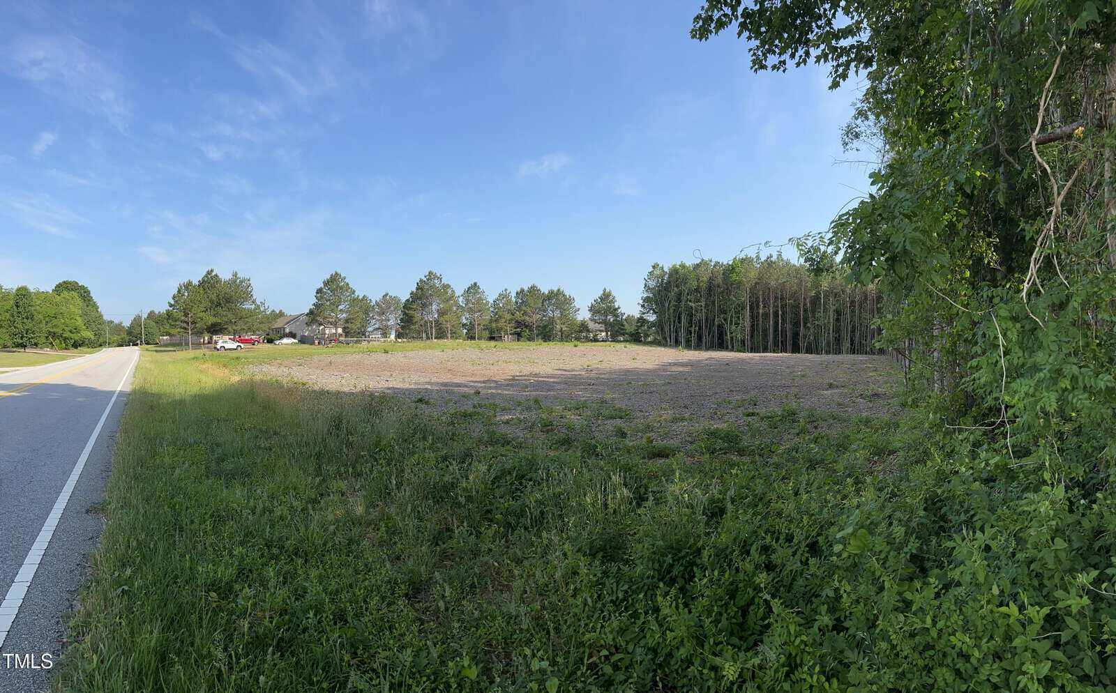 A wide, cleared lot bordered by grass and trees sits next to a rural road under a blue sky, with a few houses and vehicles visible in the background.