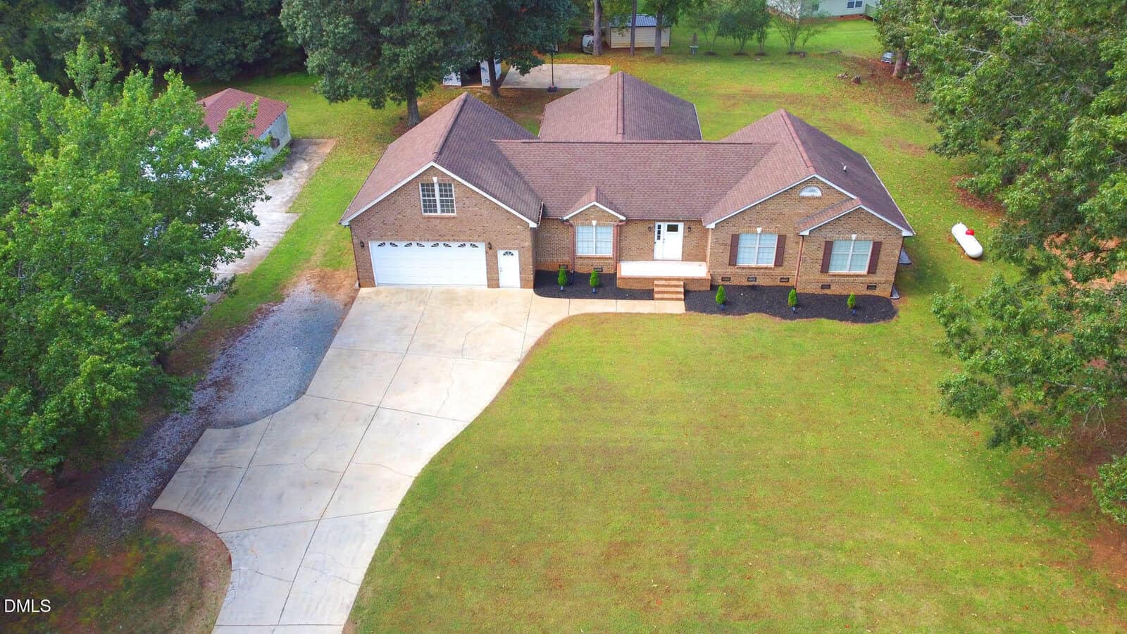 Aerial view of a single-story brick house with a two-car garage, front steps, and a large driveway. The property has a well-kept lawn, some trees, and a small shed in the backyard.