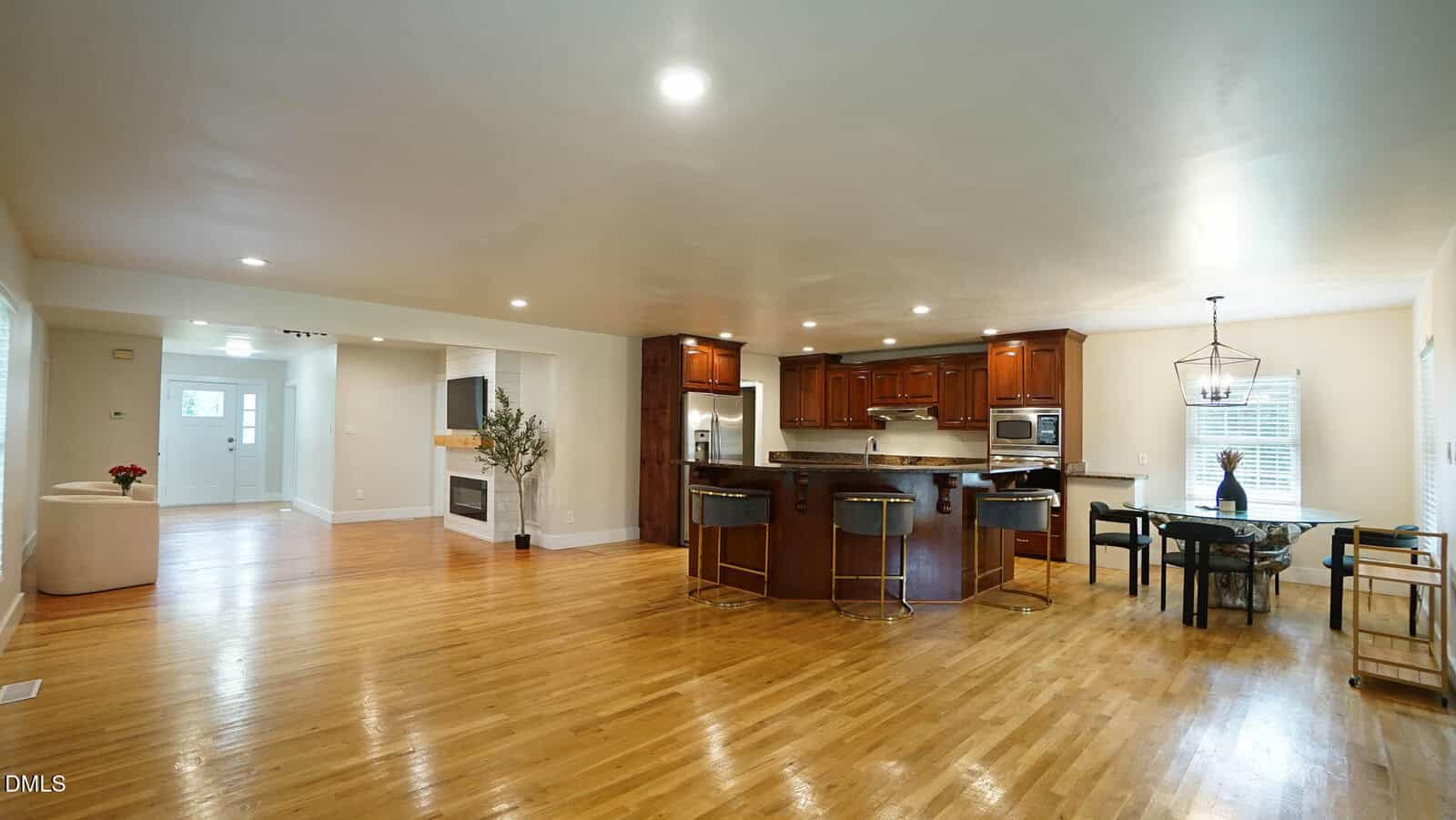 Spacious open-plan kitchen and dining area with wooden floors, a large island with bar stools, dark wood cabinets, stainless steel appliances, and a dining table near windows. Living area visible in the background.