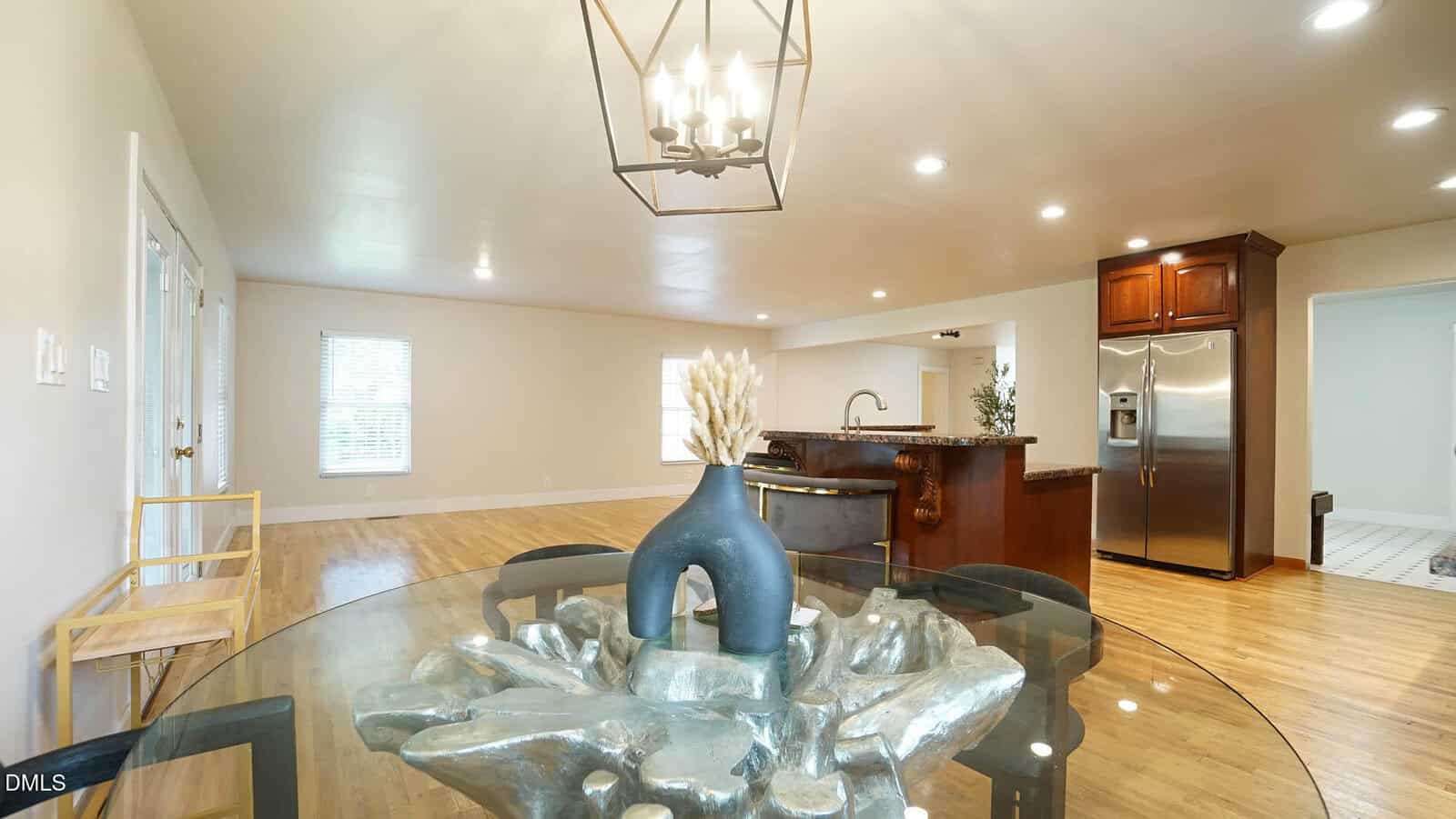 Bright open kitchen and dining area with wooden floors, round glass table topped with a vase, modern light fixture, and stainless steel appliances in the background.