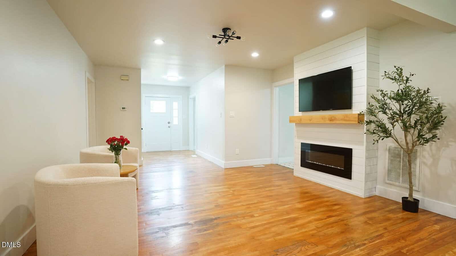 A living room with light-colored walls and wood flooring features two cream chairs with a small table and red flowers, a fireplace with a wall-mounted TV, and a potted plant; a hallway leads to a white front door.