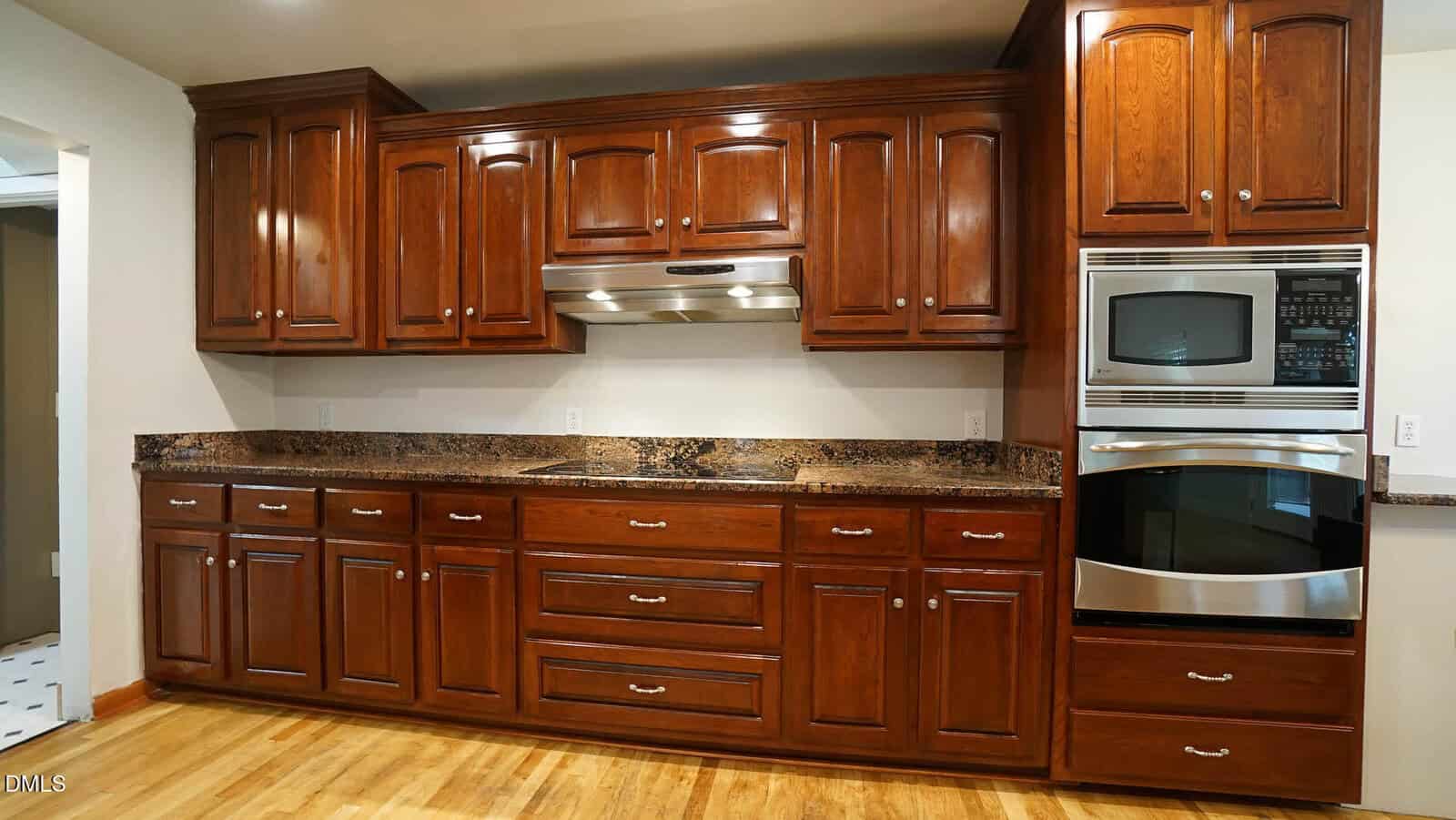 A kitchen with wooden cabinets, granite countertops, a stainless steel range hood, a built-in microwave, and an oven. The floor is hardwood, and the walls are neutral-colored. No visible appliances or decorations.