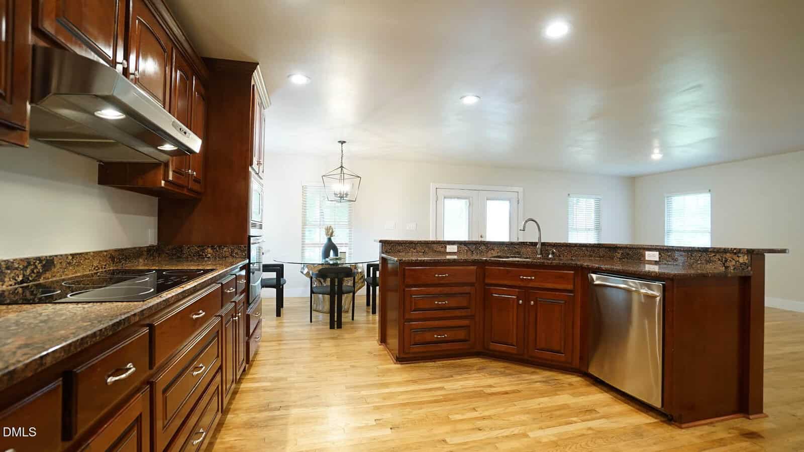 Modern kitchen with wood cabinets, granite countertops, stainless steel appliances, and hardwood floors. An open dining area with a table and chairs is visible in the background, along with large windows.