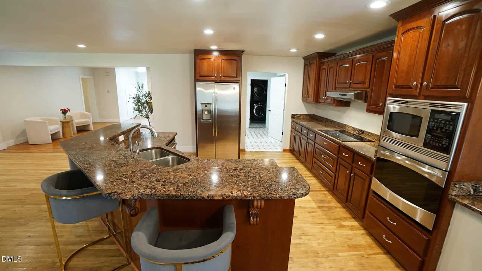 A spacious kitchen with dark wood cabinets, granite countertops, stainless steel appliances, a double oven, and a large island with two velvet barstools. Hardwood floors and recessed lighting are visible.