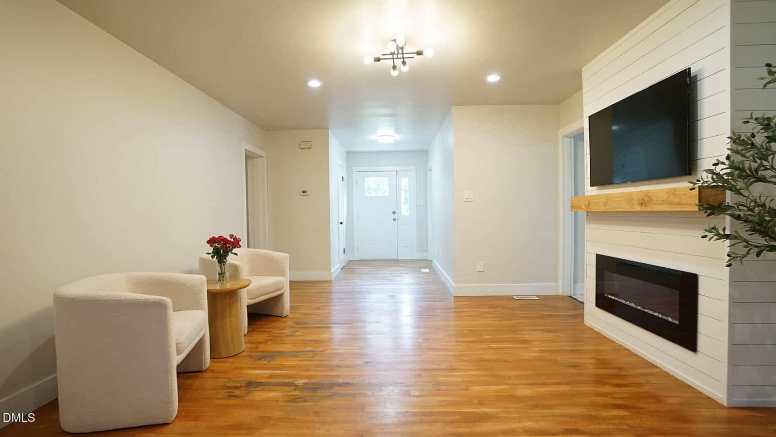 A bright hallway with wood flooring, two white chairs and a small round table with flowers on the left, and a wall-mounted TV above a fireplace on the right. Doors and a white front door are visible in the background.