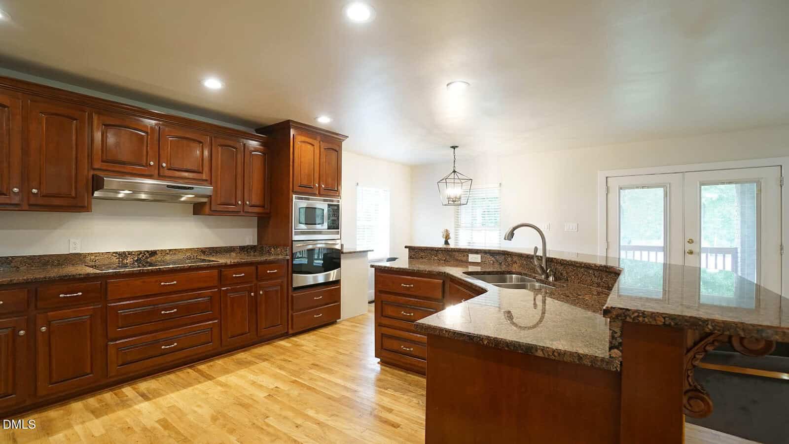 A spacious kitchen with wooden cabinets, granite countertops, double wall ovens, a built-in cooktop, a sink on a curved island, hardwood floors, recessed lighting, and double glass doors leading outside.
