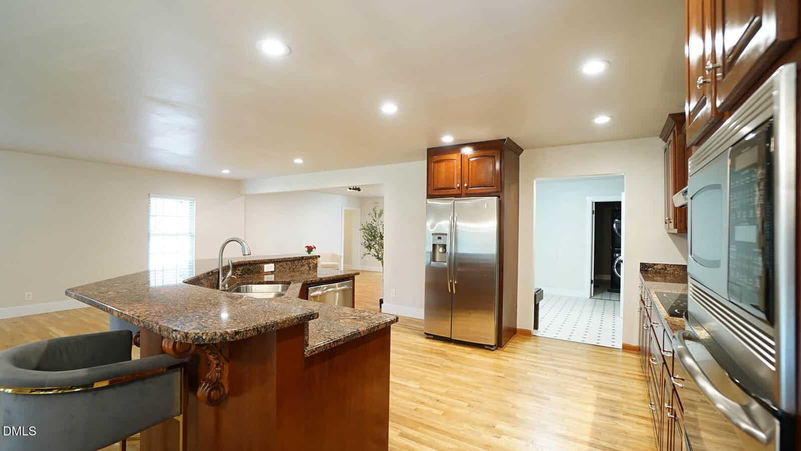 Spacious kitchen with granite countertops, stainless steel appliances, wooden cabinets, island with sink and bar seating, and light wood flooring. A doorway leads to a tiled room.