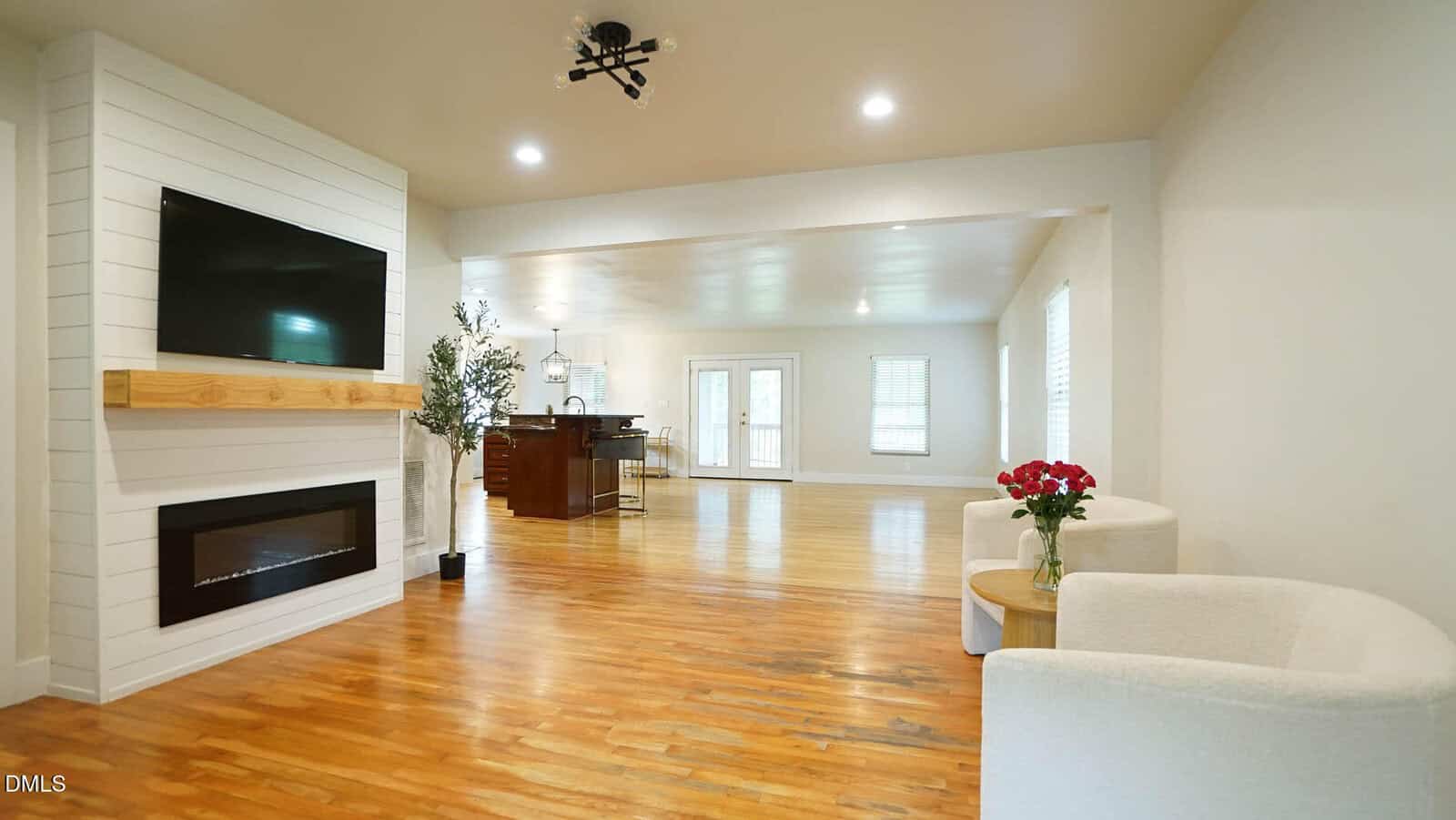 A spacious living area with hardwood floors, a wall-mounted TV above a fireplace, two white chairs with a small round table and flowers, and a kitchen area visible in the background.